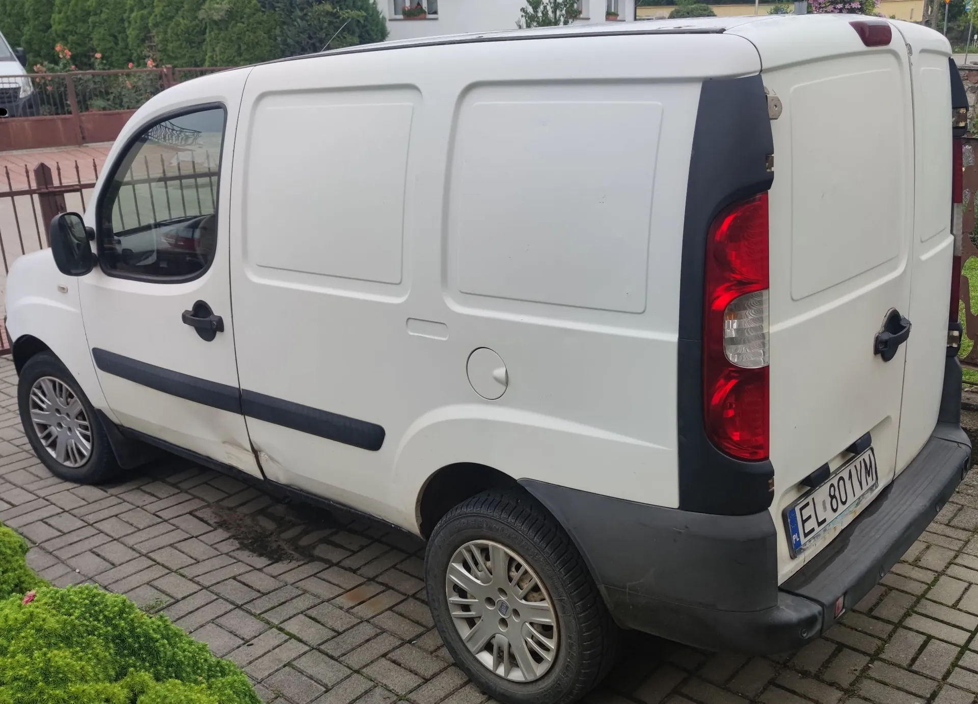 Biały samochód dostawczy typu furgon zaparkowany na kostce brukowej. Widoczny jest bok i tył pojazdu. Samochód ma czarne listwy boczne, czarne klamki i tylne światła w pionowym układzie. Tablica rejestracyjna ma numer EL 801VM.