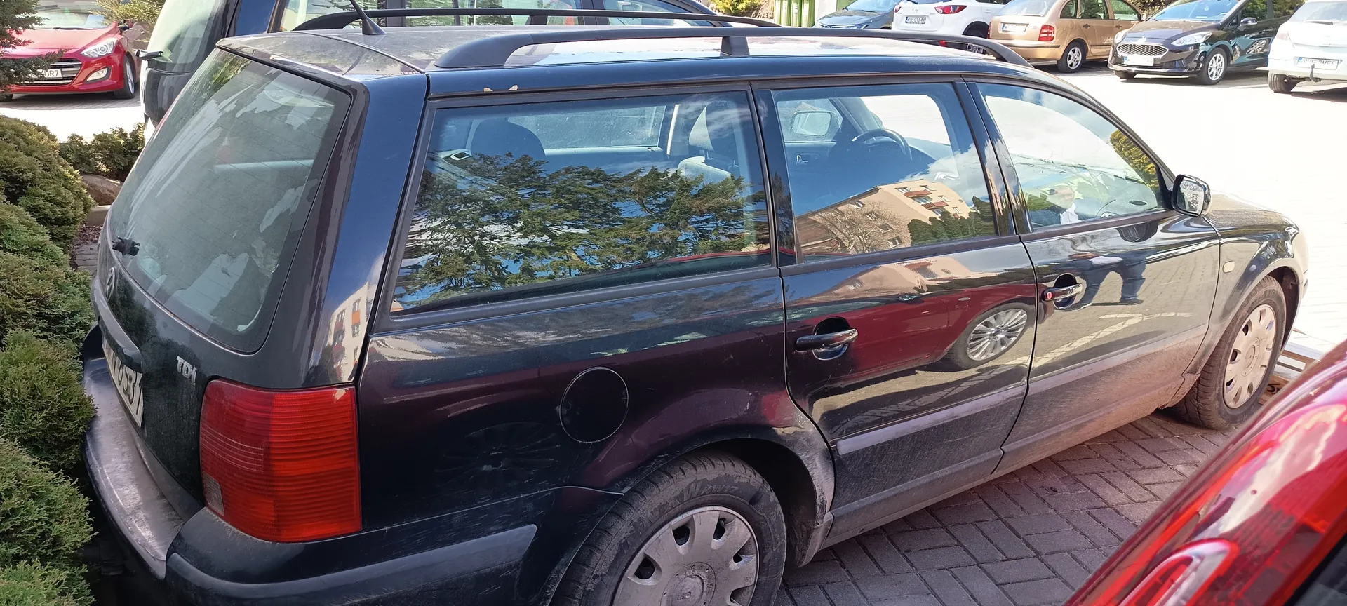 Ciemny samochód typu kombi zaparkowany na brukowanej powierzchni. Widoczne są tylna część pojazdu z czerwonymi światłami, tablicą rejestracyjną z częściowo czytelnym numerem, dachowymi prętami oraz odbiciami drzew i budynków w szybach. W tle stoją inne samochody: czerwony, biały, brązowy i czarny. Po lewej stronie obrazu rosną krzewy. Samochód ma srebrne felgi i widoczne ślady brudu na karoserii.