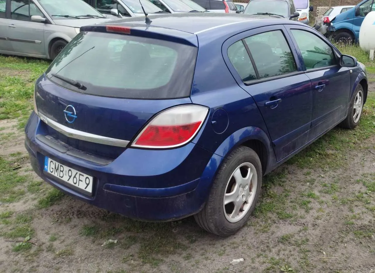 Niebieski samochód osobowy marki Opel, widok z tyłu. Na tablicy rejestracyjnej widoczny napis GMB 96F9. Samochód zaparkowany na terenie z trawą i żwirem, w tle inne pojazdy. Widoczne są czerwone światła tylnego, logo marki oraz antena na dachu.
