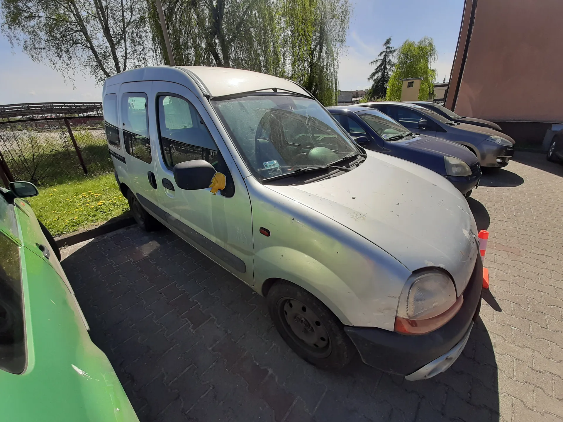 Srebrny furgonetka zaparkowana na brukowanym placu, z innymi samochodami (niebieskim i szarym) w tle. Po lewej stronie widoczny fragment zielonego samochodu. Przy przodzie furgonetki stoją pomarańczowe stożki. Tło obejmuje trawnik z kwiatami, drzewa, metalowe ogrodzenie oraz budynki. Na lusterku bocznym furgonetki znajduje się żółty przedmiot. Niebo jest jasne, słoneczna pogoda.