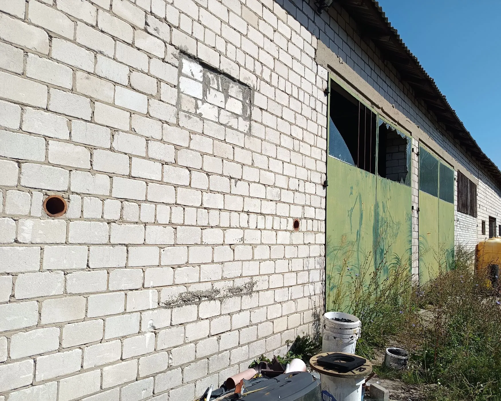 Ściana budynku wykonana z białych cegieł z widocznymi ubytkami i dziurami. Po prawej stronie znajdują się duże, zielone, metalowe drzwi garażowe, z których część jest uszkodzona i odsłania ciemne wnętrze. Przed budynkiem rośnie wysoka, dzika trawa i znajdują się różne odpady, w tym białe plastikowe wiadra.
