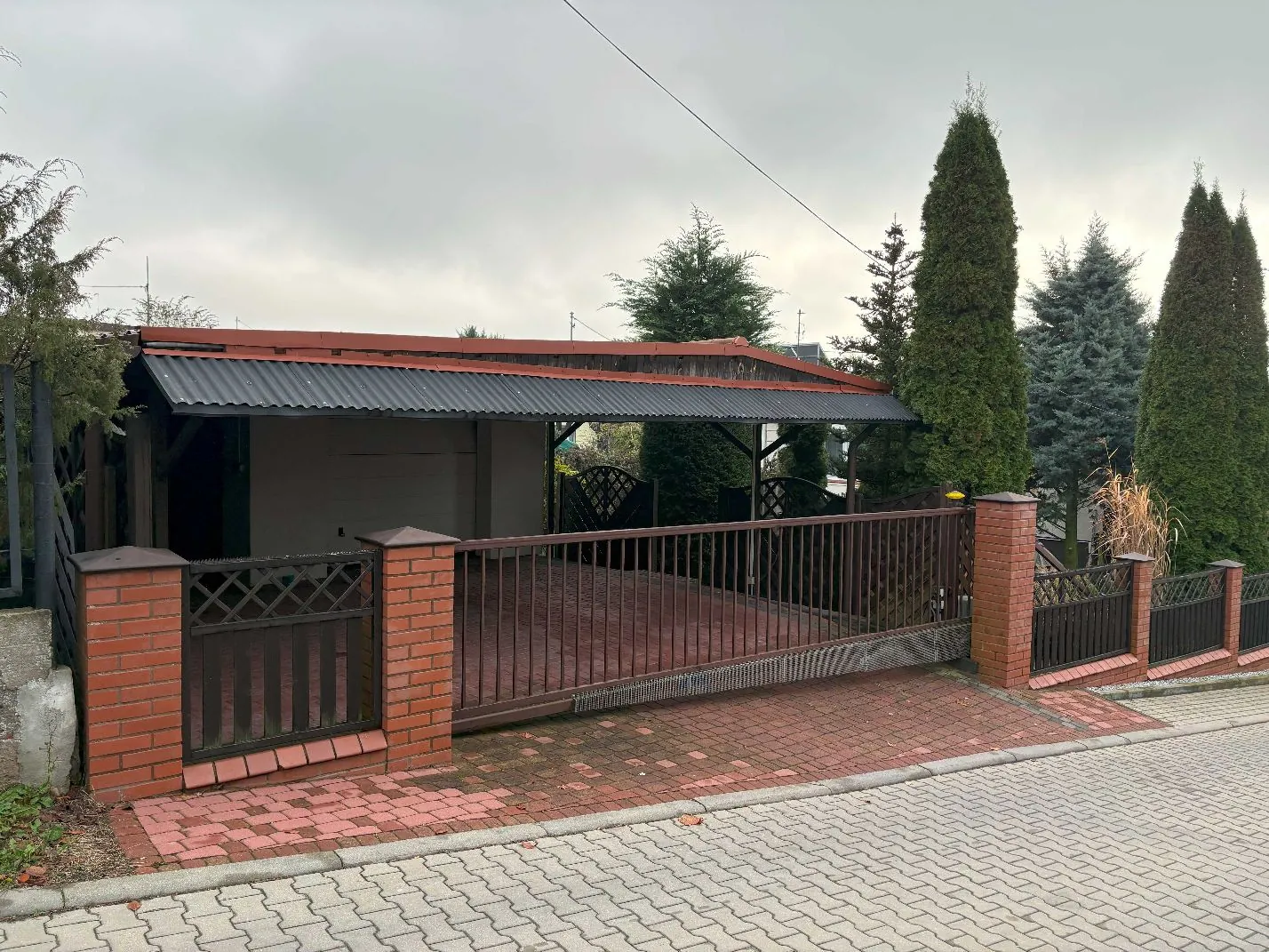 Zdjęcie przedstawia podjazd i wjazd do posesji. W centrum znajduje się brązowa, metalowa brama przesuwna z pionowymi szczeblami, osadzona między słupkami z czerwonej cegły. Za bramą widoczna jest wiata z ciemnym, falistym dachem i brązową konstrukcją. Po prawej stronie rosną wysokie, smukłe drzewa iglaste (tuje). Podjazd oraz chodnik przed bramą są wyłożone kostką brukową w kolorach szarym i czerwonym. Niebo jest zachmurzone.
