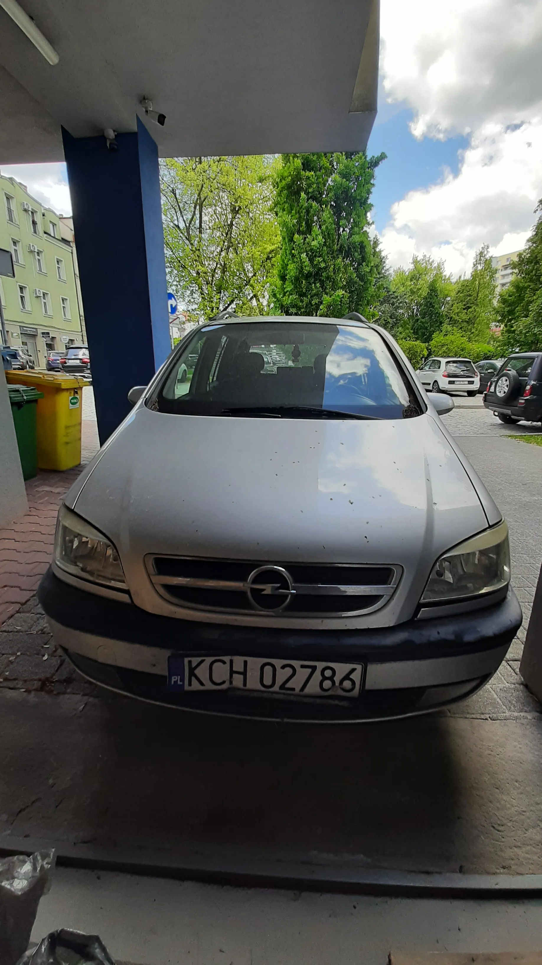 Srebrny samochód marki Opel zaparkowany na brukowanej powierzchni. Widoczna tablica rejestracyjna KCH 02786 z oznaczeniem PL. W tle inne pojazdy, drzewa, budynki, niebieska konstrukcja oraz żółte i zielone pojemniki na śmieci. Niebo częściowo zachmurzone.