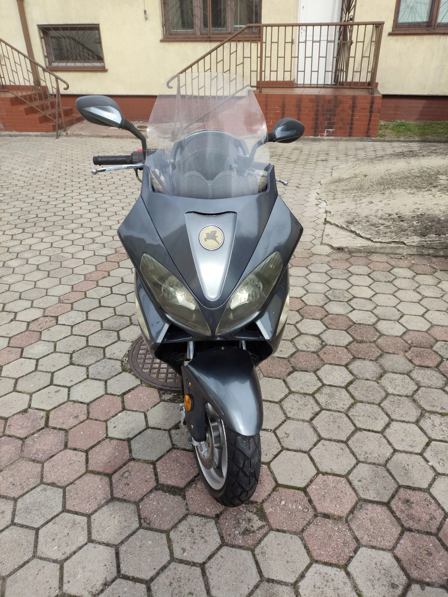 Widok z przodu na ciemnoszary motocykl zaparkowany na kostce brukowej o sześciokątnym kształcie. Motocykl posiada przezroczystą szybę ochronną, dwa reflektory oraz lusterka boczne. W tle widoczny jest fragment jasnego budynku z oknami i metalową barierką schodów.
