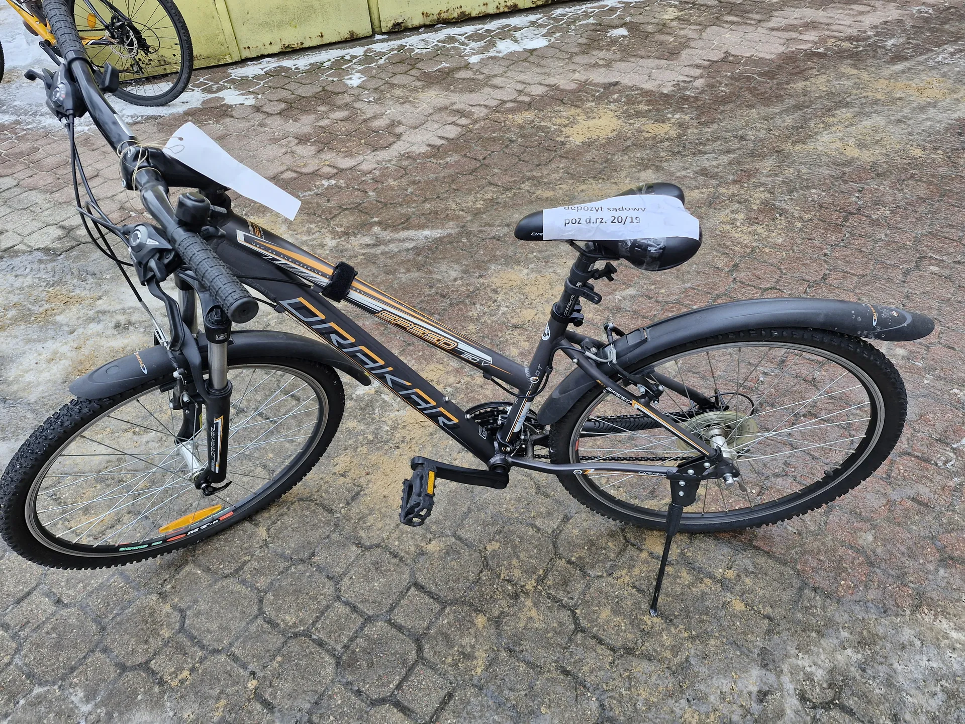 Czarny rower z pomarańczowymi akcentami marki DRAKAR stojący na brukowanej powierzchni. Na siedzeniu widoczny biały napis: 'depozyt sądowy poz d.rz. 20/19'. W tle fragment żółtej ściany i części innego roweru. Na kierownicy umocowany biały kartonik. Rower oparty o podstawę, z widocznymi oponami, kierownicą i mechanizmem przerzutek.
