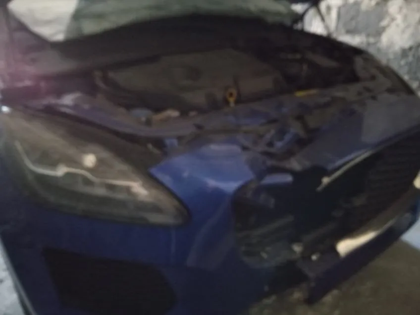 Niebieski samochód z otwartą maską, uszkodzonym przodem (zderzak, atrapa chłodnicy) i widocznym kompartem silnika. Tło stanowi kamienna ściana.