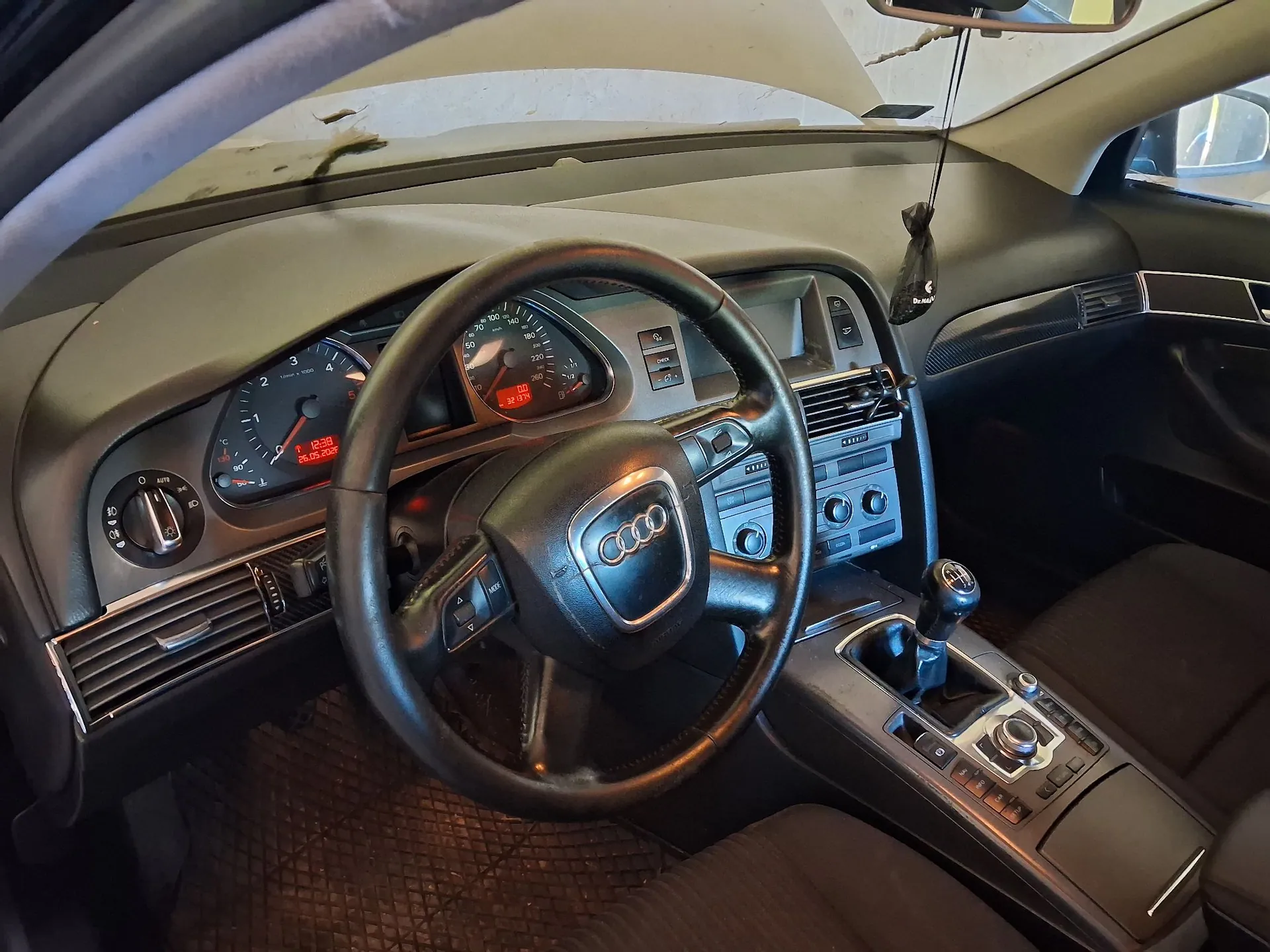 Wnętrze samochodu marki Audi, widok z perspektywy kierowcy. Widoczna jest czarna skórzana kierownica z logo Audi w centrum, deska rozdzielcza z analogowymi zegarami prędkościomierza i obrotomierza, konsola środkowa z radiem i lewarkiem zmiany biegów oraz czarna tapicerka foteli.