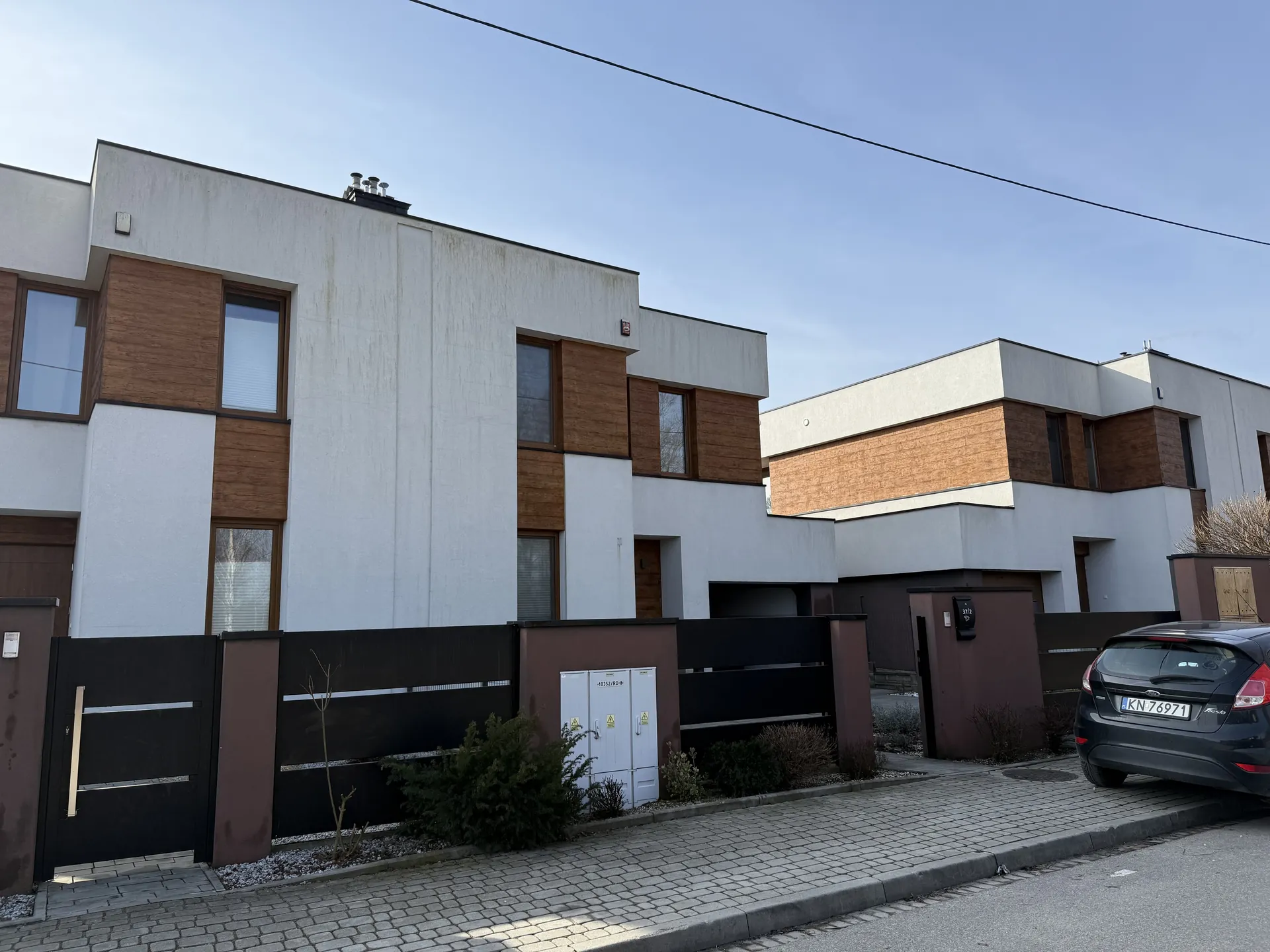Nowoczesne budynki mieszkalne z białymi i brązowymi elewacjami, z oknami o brązowych ramach. Przed budynkami czarne ogrodzenie z bramą, biały szafka elektryczna z żółtymi znakami ostrzegawczymi. Na prawo czarny samochód osobowy z tablicą rejestracyjną KN 76971. Chodnik z kostki brukowej, droga asfaltowa, jasne niebo z przewodami energetycznymi. Rośliny i krzewy przy ogrodzeniu.