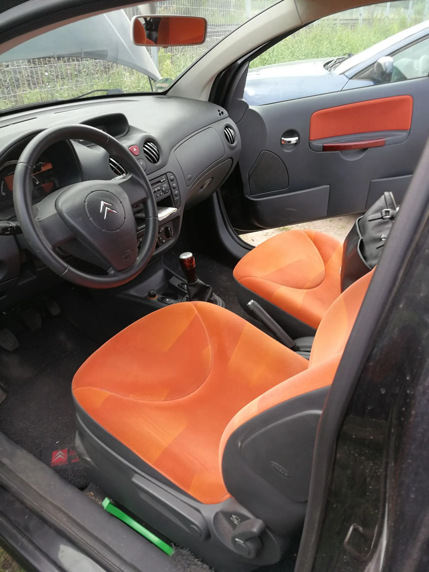 Wnętrze samochodu widziane z otwartych drzwi pasażera. Widoczna jest kierownica z logo Citroëna, deska rozdzielcza, dwa fotele z pomarańczową tapicerką oraz czarne panele boczne drzwi z pomarańczowym akcentem. W tle widać fragment lusterka wstecznego oraz inny niebieski samochód zaparkowany obok.