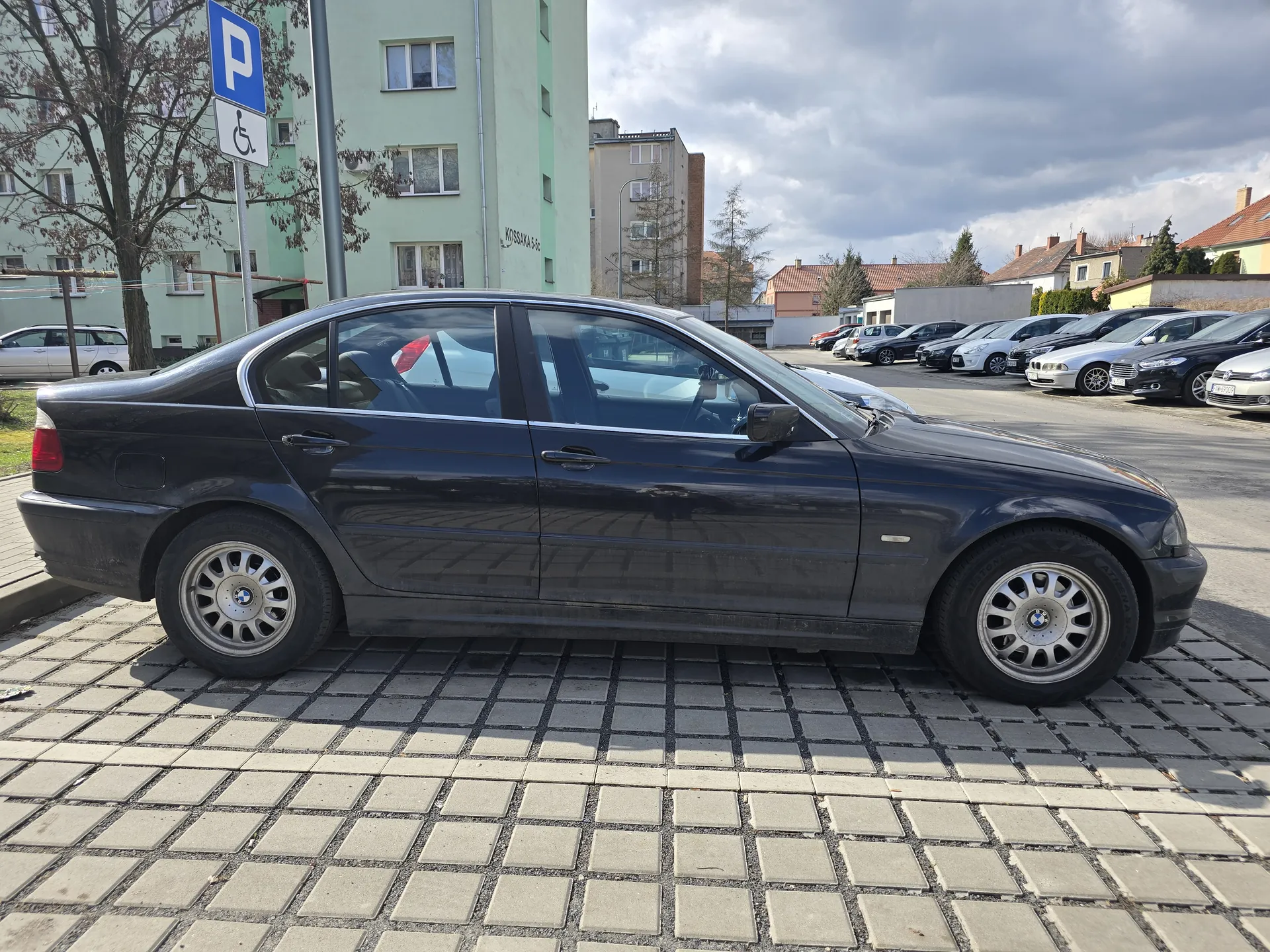 Czarny samochód marki BMW zaparkowany na wybrukowanym placu parkingowym. W tle niebieski znak parkingowy z literą 'P' i symbolem niepełnosprawności, zielony blok mieszkalny z napisem 'KOSAKA 5-6', inne zaparkowane pojazdy, drzewa bez liści oraz częściowo zachmurzone niebo. Na pierwszym planie chodnik z kwadratowych płyt.