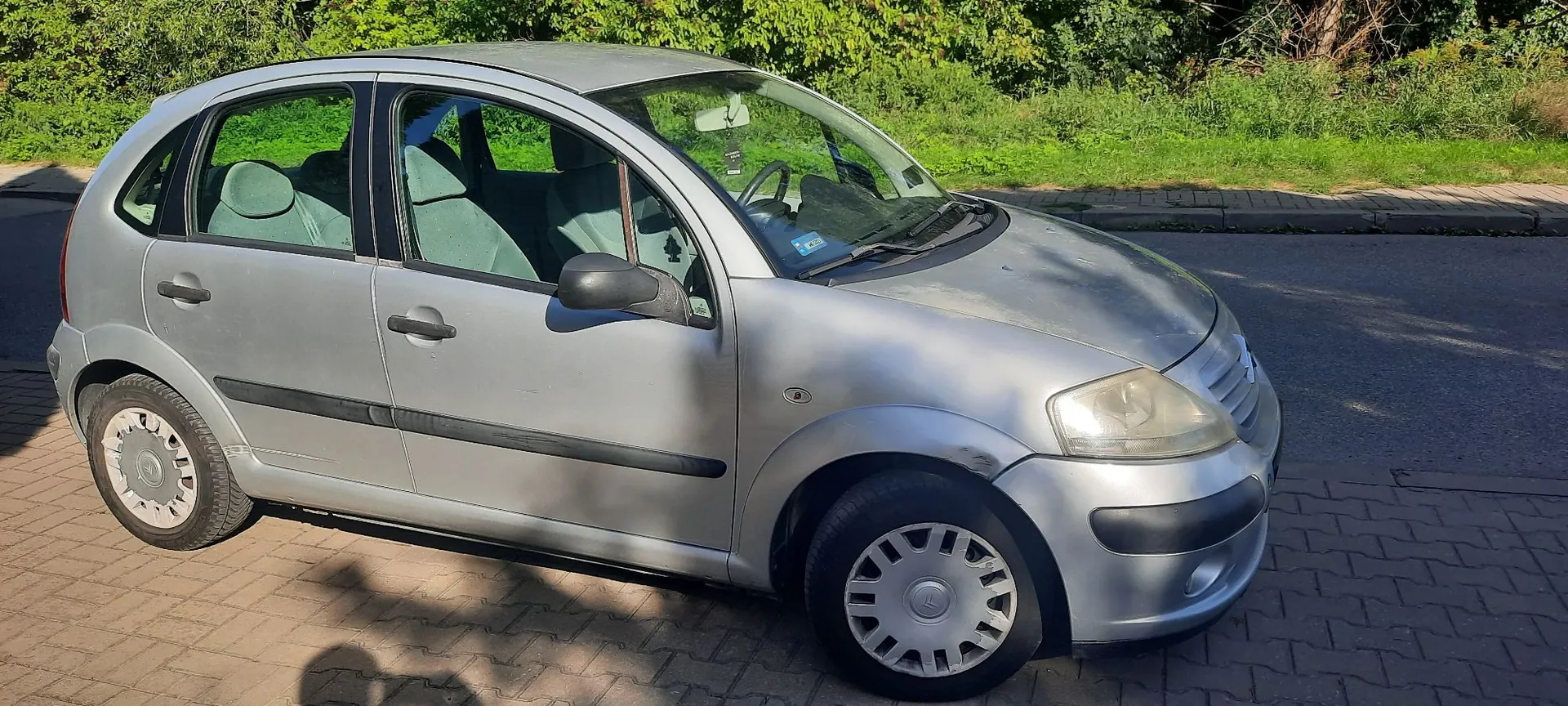 Srebrny, pięciodrzwiowy samochód osobowy marki Renault Clio drugiej generacji, zaparkowany na kostce brukowej przy krawężniku. Auto jest widoczne z profilu od strony pasażera. W tle widać zielone krzewy i trawę.