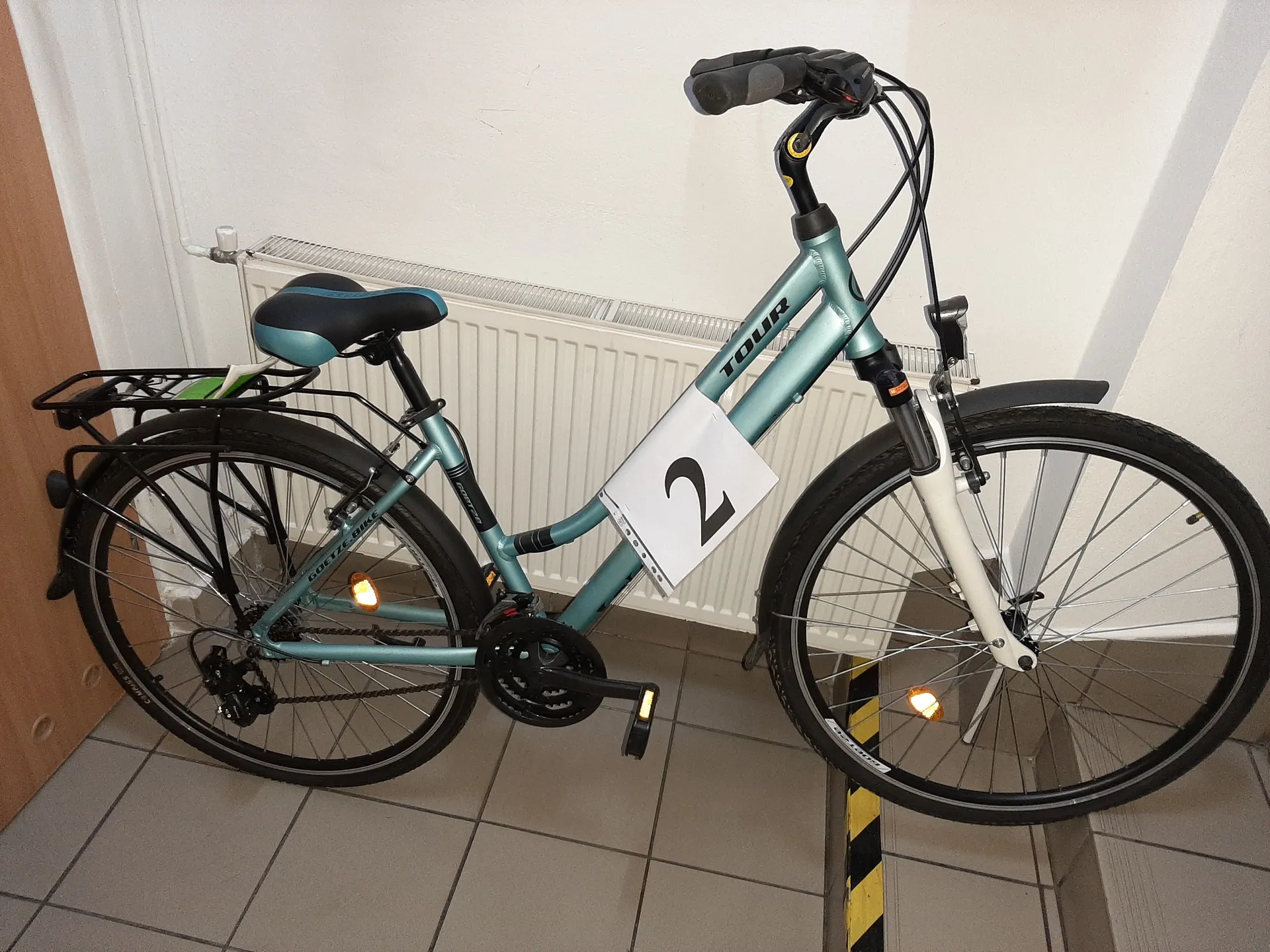 Niebiesko-zielony rower z napisem 'TOUR' na ramie, z białą tabliczką z czarną cyfrą '2' przymocowaną do ramy. Rower ma czarno-niebieskie siedzisko, bagażnik tylny z zielonym przedmiotem, włączonymi pomarańczowymi reflektorami oraz czarnymi oponami. Znajduje się w pomieszczeniu na tle białego grzejnika, białej ściany i podłogi z szarymi płytkami. Po lewej stronie widoczna jest brązowa drzwi.