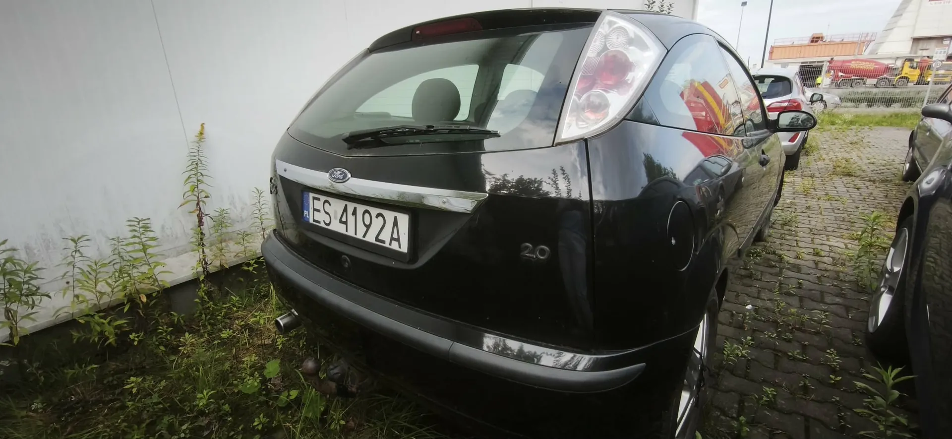Tylny widok czarnego samochodu marki Ford Focus z polską tablicą rejestracyjną ES 4192A. Na klapie bagażnika widnieje oznaczenie 2.0. Samochód zaparkowany jest na kostce brukowej obok białej ściany i innych pojazdów.