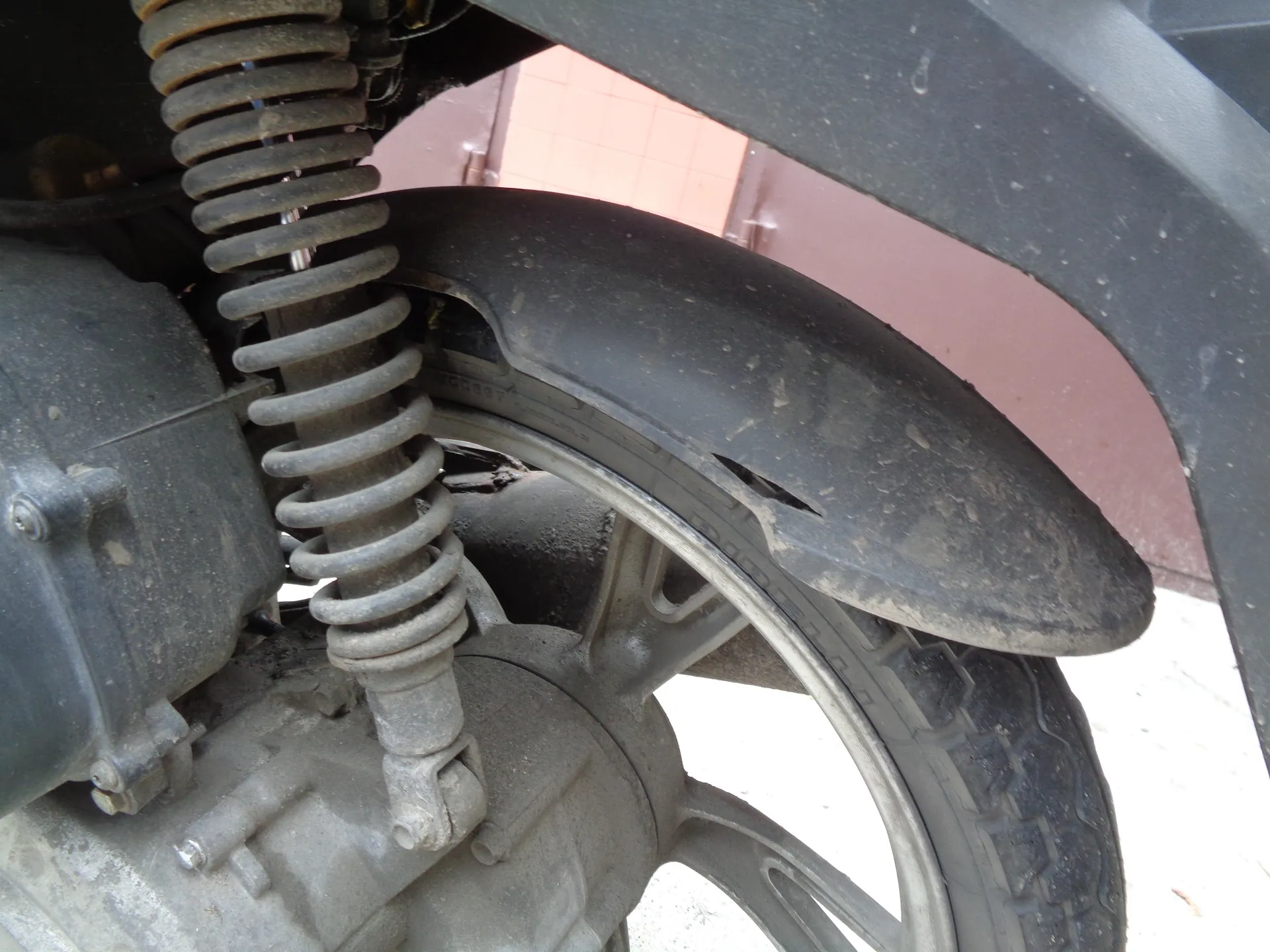 Zbliżenie na tylne koło i zawieszenie motocykla. Widoczny jest amortyzator z metalową sprężyną, czarny błotnik z widocznym pęknięciem oraz fragment opony i felgi. Całość jest pokryta warstwą kurzu i brudu.