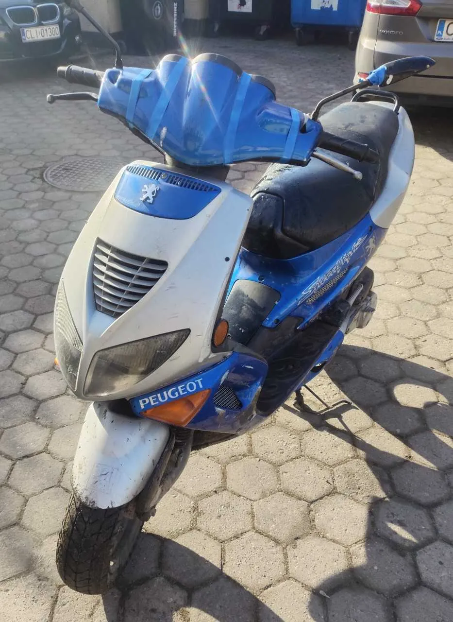 Niebiesko-biały skuter marki PEUGEOT zaparkowany na brukowanej powierzchni z sześciokątnymi płytami. Na przedniej części skutera widoczny jest logo Peugeota oraz napis "PEUGEOT" na niebieskim tle. Skuter posiada czarną siedzisko i niebieską osłonę przednią z taśmą. W tle znajdują się samochody: czarny BMW z tablicą rejestracyjną "CL 01300" oraz beżowy samochód z częściowo widoczną tablicą "PL 0...". W oddali widoczne są niebieskie i czarne pojemniki na śmieci.
