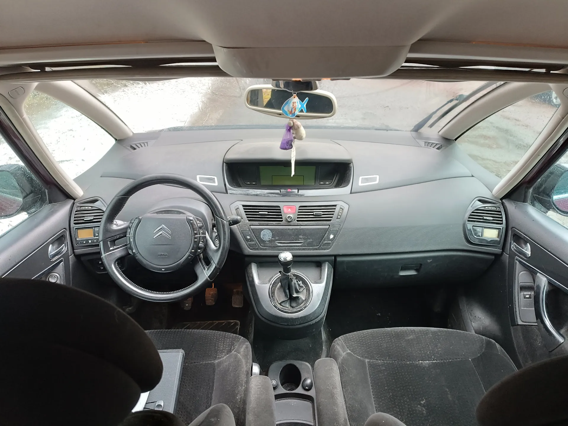 Wnętrze samochodu Citroën z kierownicą z logo, szarą deską rozdzielczą z wyświetlaczem, czarnymi siedzeniami tapicerowanymi tkaniną i manualną skrzynią biegów. W lusterku wstecznym wiszą ozdoby: niebieska rybka i fioletowy przedmiot. Poza oknami widoczny śnieg.