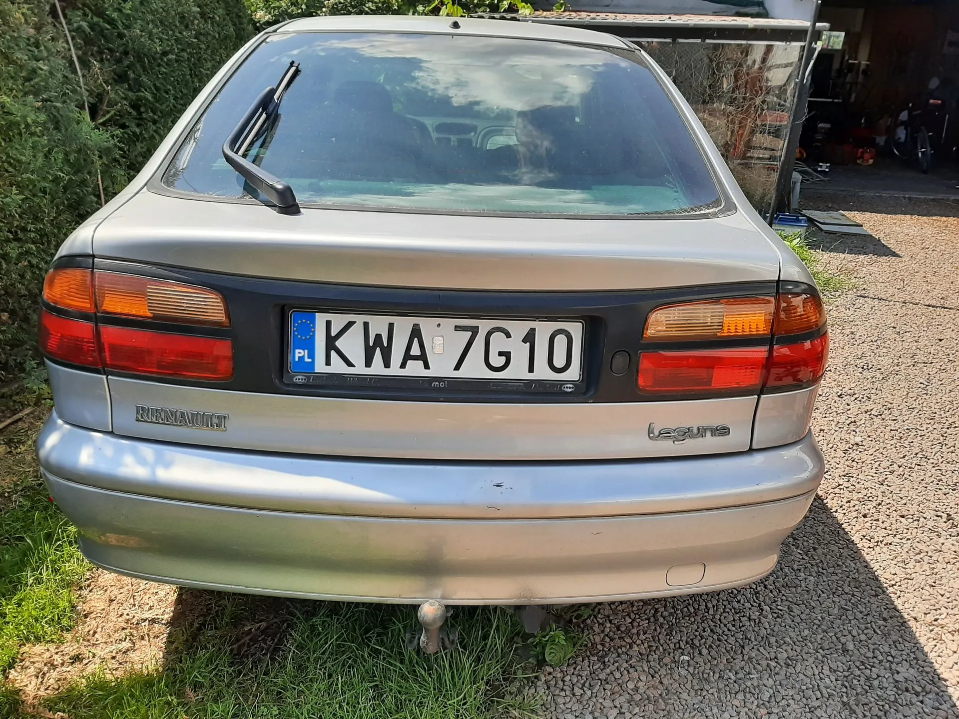 Tylny widok srebrnego samochodu marki Renault Laguna. Na klapie bagażnika znajduje się polska tablica rejestracyjna o numerze KWA 7G10. Po lewej stronie widnieje napis RENAULT, a po prawej Laguna. Samochód zaparkowany jest na żwirowym podjeździe obok zieleni.