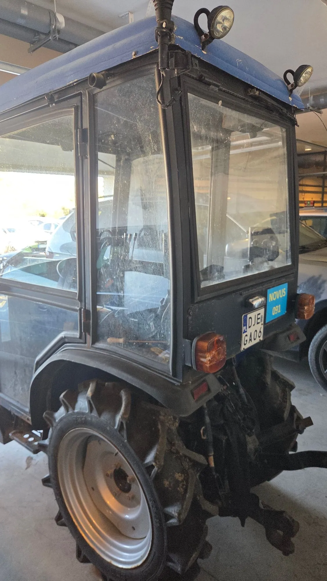 Niebieski traktor z przeszkloną kabiną, zaparkowany w garażu. Na tylnej szybie widoczna jest niebieska naklejka z napisem 'NOVUS 091' oraz tablica rejestracyjna o numerze 'DJE GA06'. Pojazd posiada duże, błotniste koła z charakterystycznym bieżnikiem oraz reflektory zamontowane na dachu.