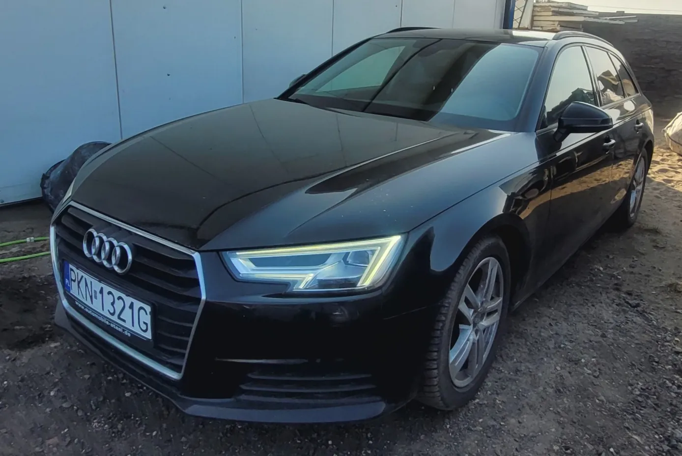 Czarny samochód Audi Avant zaparkowany na nierównym, żwirowym podłożu. Widoczna przód pojazdu z charakterystyczną atrapą chłodnicy z logo Audi, reflektorem LED oraz tablicą rejestracyjną PKN 1321G. W tle błękitna ściana i materiały budowlane. Pod tablicą rejestracyjną widoczny napis www.automobile-steel.de.