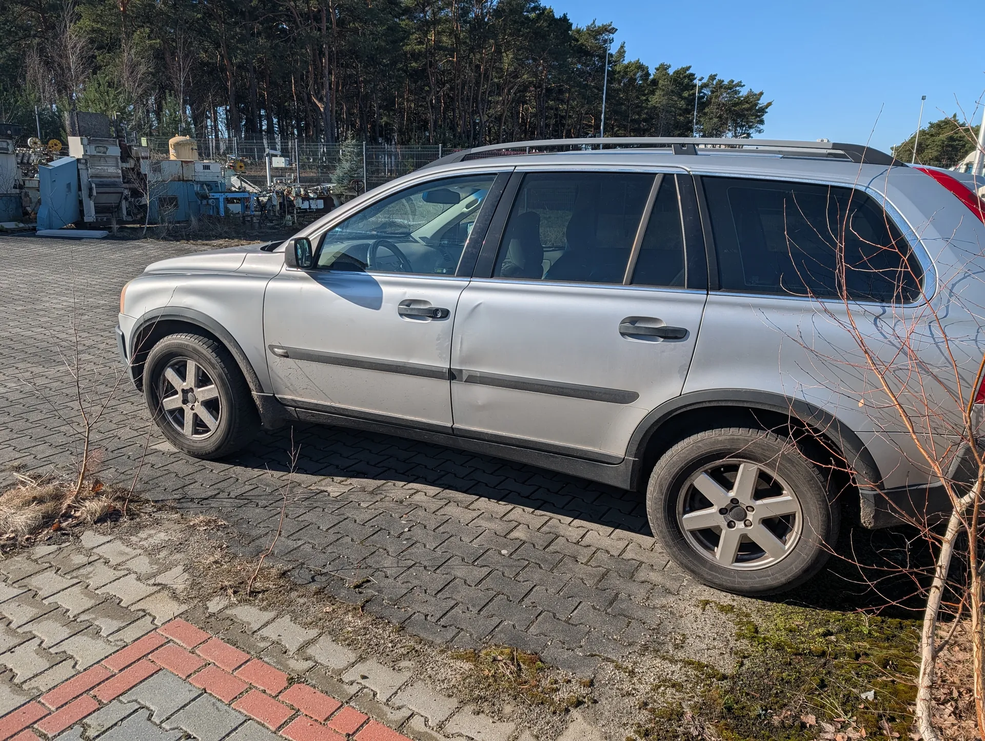 Srebrny SUV zaparkowany na powierzchni z kostki brukowej w kolorach szarym i czerwonym. Po lewej stronie widoczne są suche krzewy, a w tle las iglasty, ogrodzenie i maszyny przemysłowe. Niebo jest bezchmurne i błękitne. Samochód ma czarne listwy boczne, dachowy bagażnik i srebrne felgi. Wnętrze pojazdu jest częściowo widoczne przez szyby.