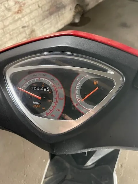 Zbliżenie na deski rozdzielcze skutera z czarnym i czerwonym obudowaniem. Po lewej stronie znajduje się wskaźnik prędkości z zakresem od 0 do 80 km/h, na wyświetlaczu cyfrowym widoczny przebieg 004494 km. Poniżej napisy 'km/h' i 'mph'. Po prawej stronie wskaźnik paliwa z ikoną zbiornika paliwowego i czerwoną strzałką. Tło przedstawia fragment betonowej ściany i podłogi.