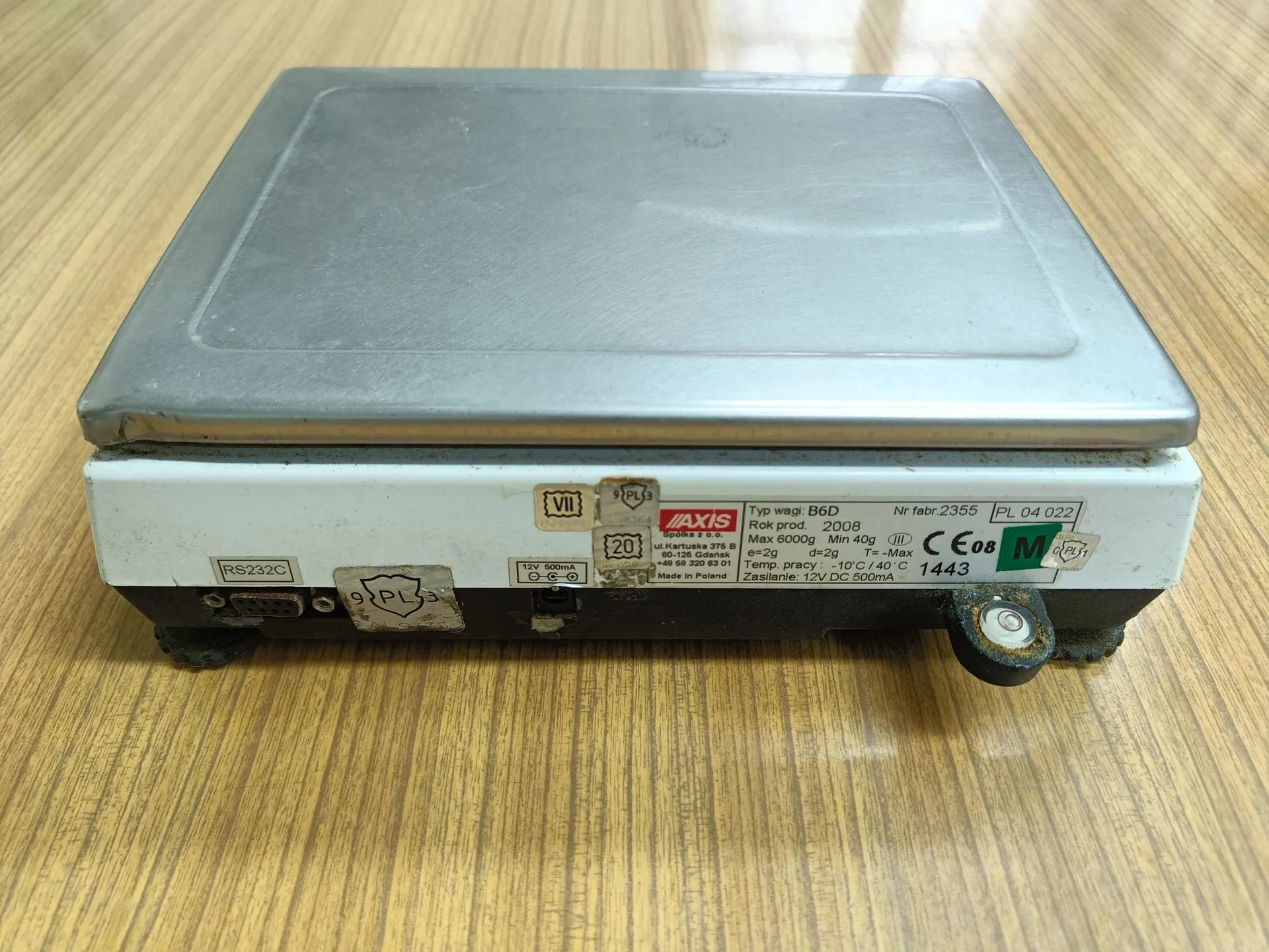 Srebrna waga elektroniczna z płaskim blatem, ustawiona na drewnianym blacie. Na przednim panelu urządzenia znajduje się tabliczka znamionowa z tekstem: 'AXIS Spółka z o.o. ul. Lądnica 10 80-125 Gdańsk tel. +48 58 301 11 11 Made in Poland', 'Typ wagi: B6D', 'Rok prod. 2008', 'Max 6000g Min 40g', 'e=2g d=2g', 'T= -10°C / +40°C', 'Zasilanie: 12V DC 500mA', 'Nr fabr. 2355', 'PL 04 022', 'CE 08', '1443'. Po lewej stronie widoczne jest złącze RS232C oraz gniazdo zasilania 12V 500mA.