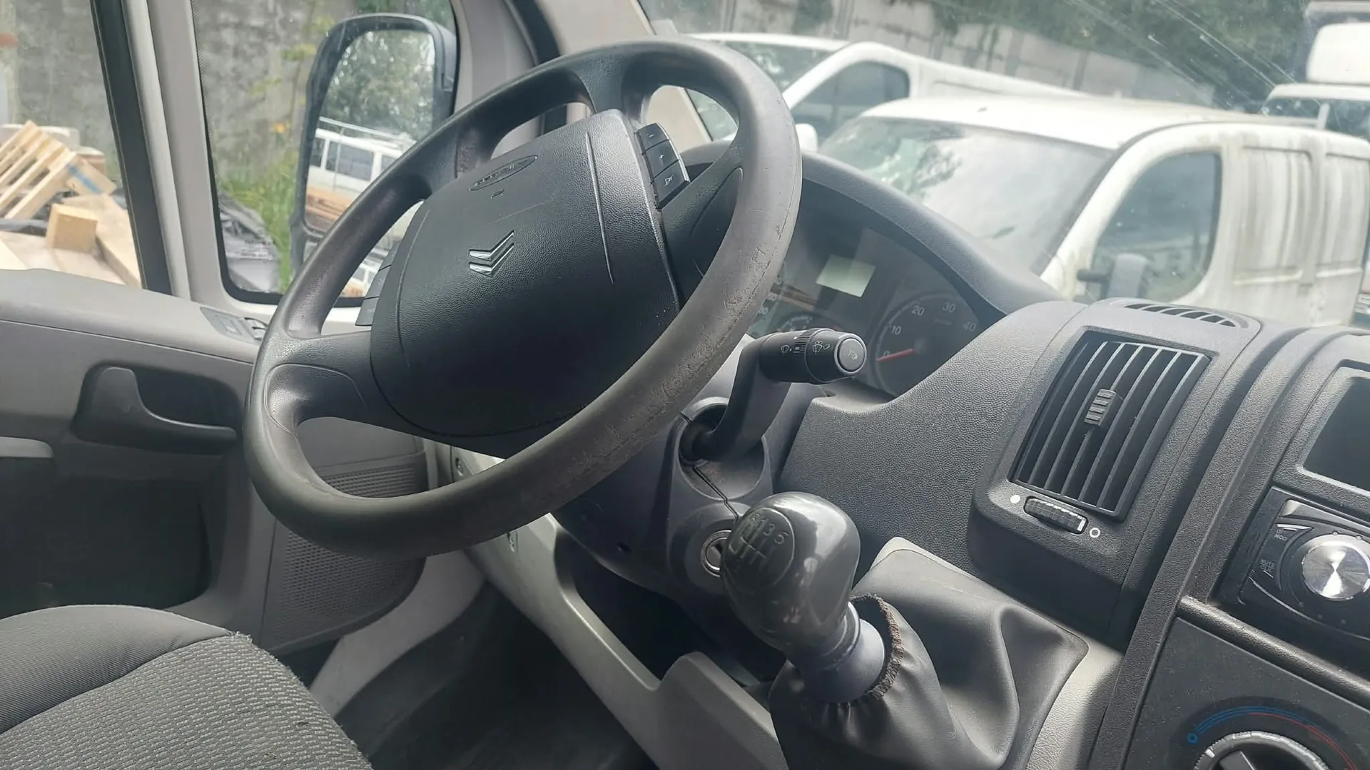 Widok z wnętrza kabiny samochodu, skupiony na kierownicy i desce rozdzielczej. Widoczna jest czarna, czteroramienna kierownica z logo marki Citroën. Za nią znajduje się panel wskaźników z prędkościomierzem. Po prawej stronie widać kratkę nawiewu oraz fragment konsoli środkowej. Przez przednią szybę i lusterko boczne widoczne są inne białe samochody dostawcze zaparkowane na zewnątrz.