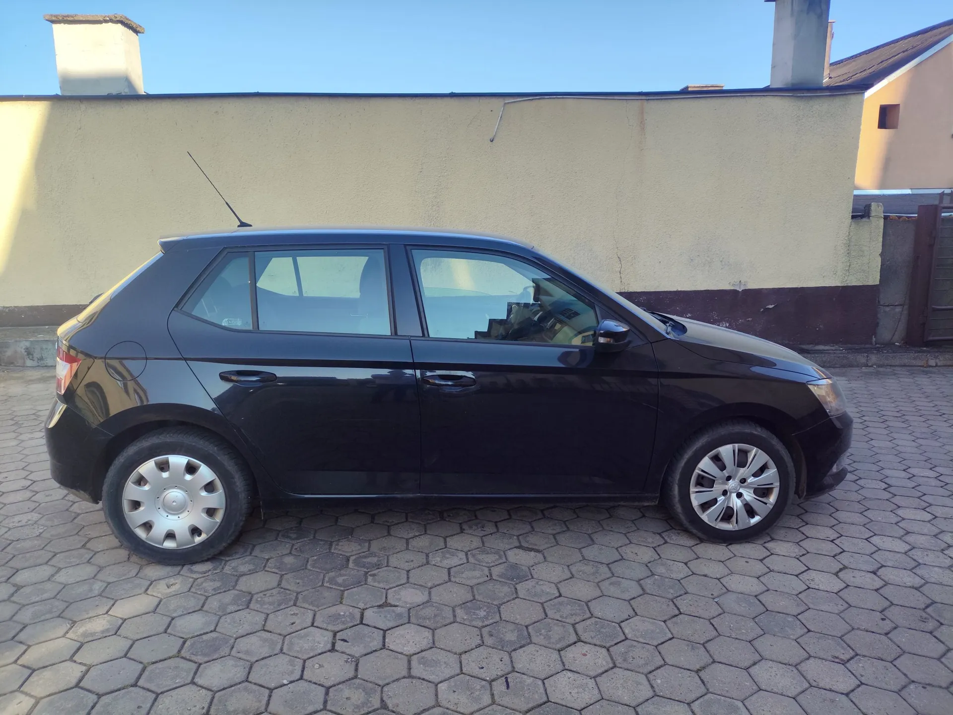 Czarny samochód osobowy typu hatchback zaparkowany bokiem na podjeździe z szarej kostki brukowej o sześciokątnym kształcie. W tle znajduje się jasna, gładka ściana budynku oraz fragmenty dachu i komina pod czystym, niebieskim niebem.