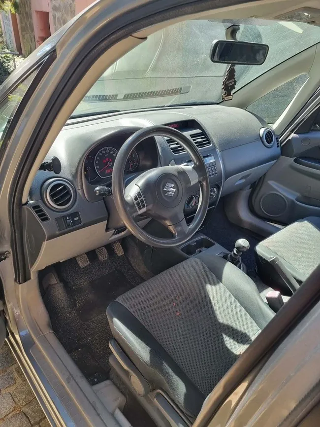 Wnętrze samochodu marki Suzuki widziane z perspektywy otwartych drzwi pasażera. Widoczna jest kierownica z logo Suzuki, deska rozdzielcza z zegarami, centralna konsola z nawiewami i radiem oraz dwa przednie fotele z ciemnej tkaniny. Na lusterku wstecznym wisi ozdoba.