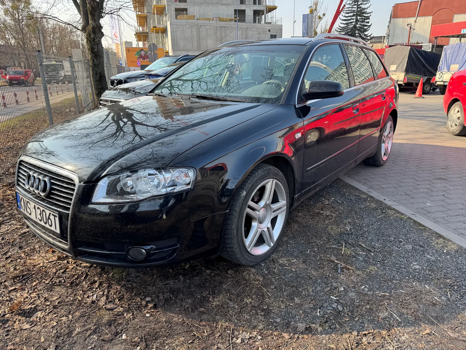 Czarny samochód Audi kombi z tablicą rejestracyjną KNS 1306T zaparkowany na żwirowym obszarze. W tle widoczne są inne pojazdy, w tym czerwony samochód i ciężarówka z pokrytą bryłą, ogrodzenie metalowe, drzewa bez liści, budowa budynku z żółtymi rusztowaniami oraz pomarańczowy stożek drogowy. Na ziemi sucha trawa i liście. Samochód ma srebrne felgi i reflektory z przodu.