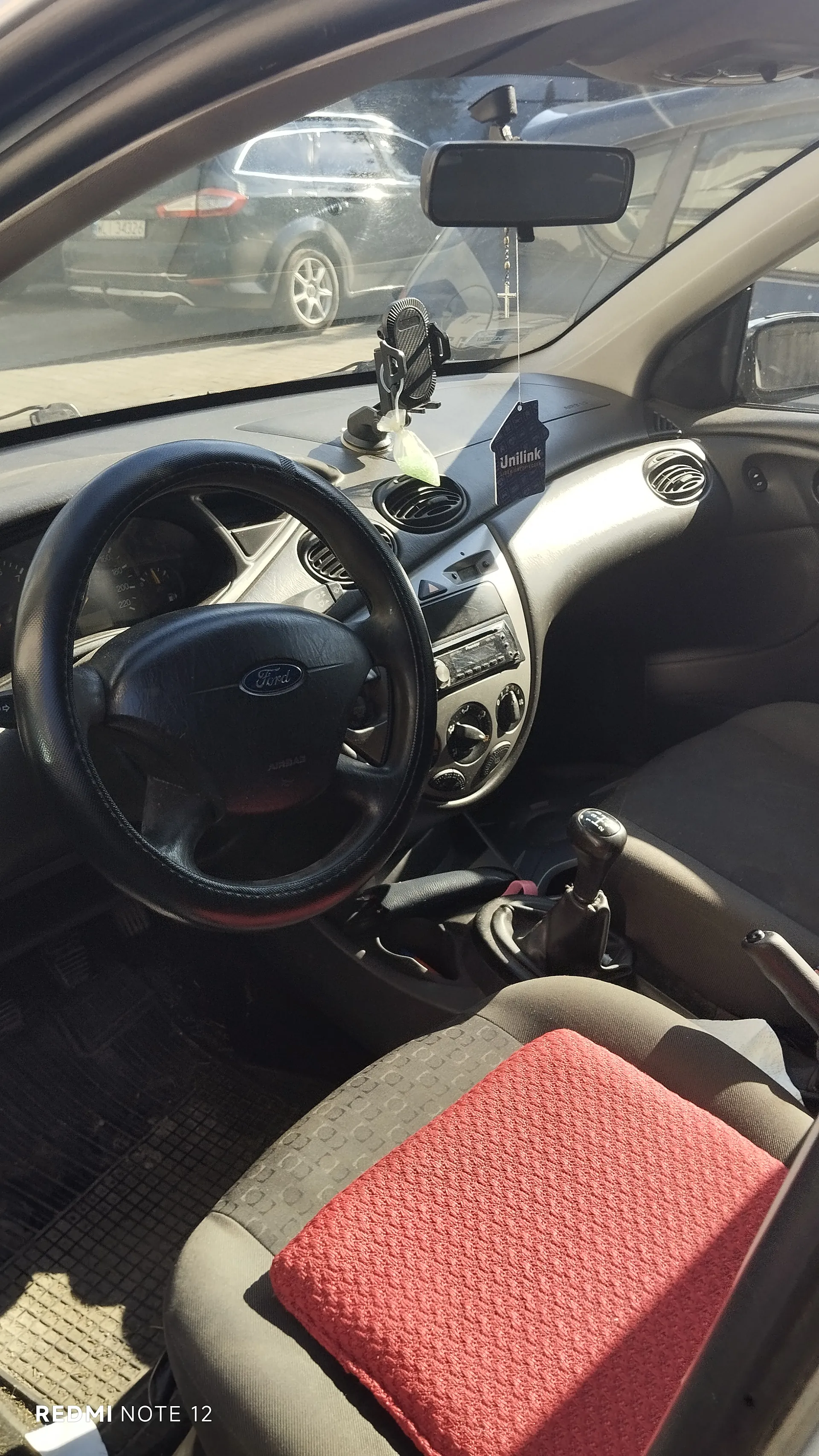 Wnętrze samochodu marki Ford. Widoczna kierownica z logo Ford, deska rozdzielcza z radioodtwarzaczem, wentylacjami i przyciskami sterującymi. Na desce rozdzielczej umieszczono wisiorek z napisem "Unilink" oraz krzyżyk wiszący na lusterku wstecznym. W tle przez przednią szybę widać zaparkowany czarny samochód. Na siedzeniu pasażera leży czerwona tkanina. W lewym dolnym rogu obrazu widoczny napis "REDMI NOTE 12".