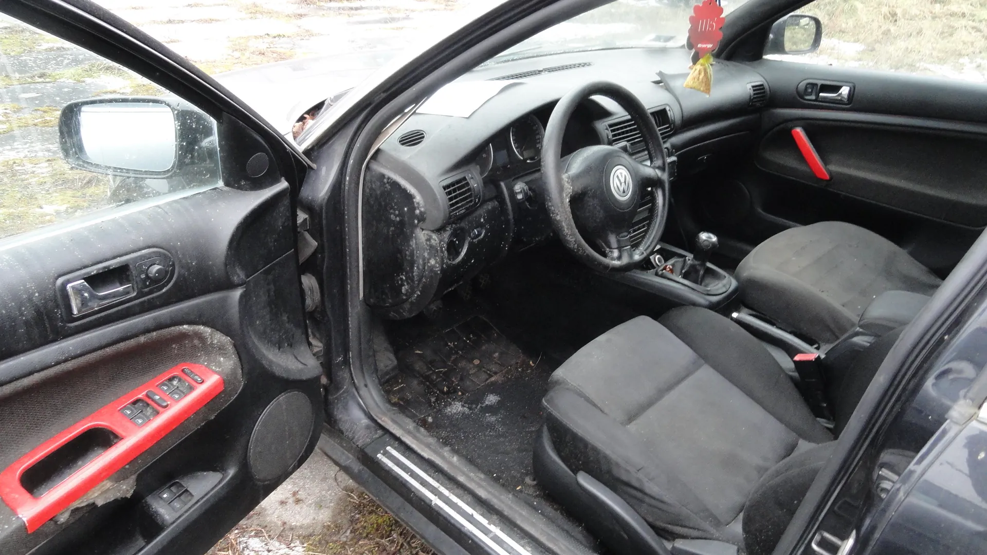 Wnętrze czarnego samochodu marki Volkswagen z otwartymi drzwiami od strony kierowcy. Widoczne są czarne siedzenia, kierownica z logo VW, deska rozdzielcza z zanieczyszczeniami oraz czerwone akcenty na uchwytach drzwiowych. Na lusterku wstecznym wiszą ozdoby: czerwony element z napisem "115" oraz żółty przedmiot. Podłoga w aucie zawiera śmieci i śnieg. Poza samochodem widoczny jest zimowy krajobraz z trawą i śniegiem.