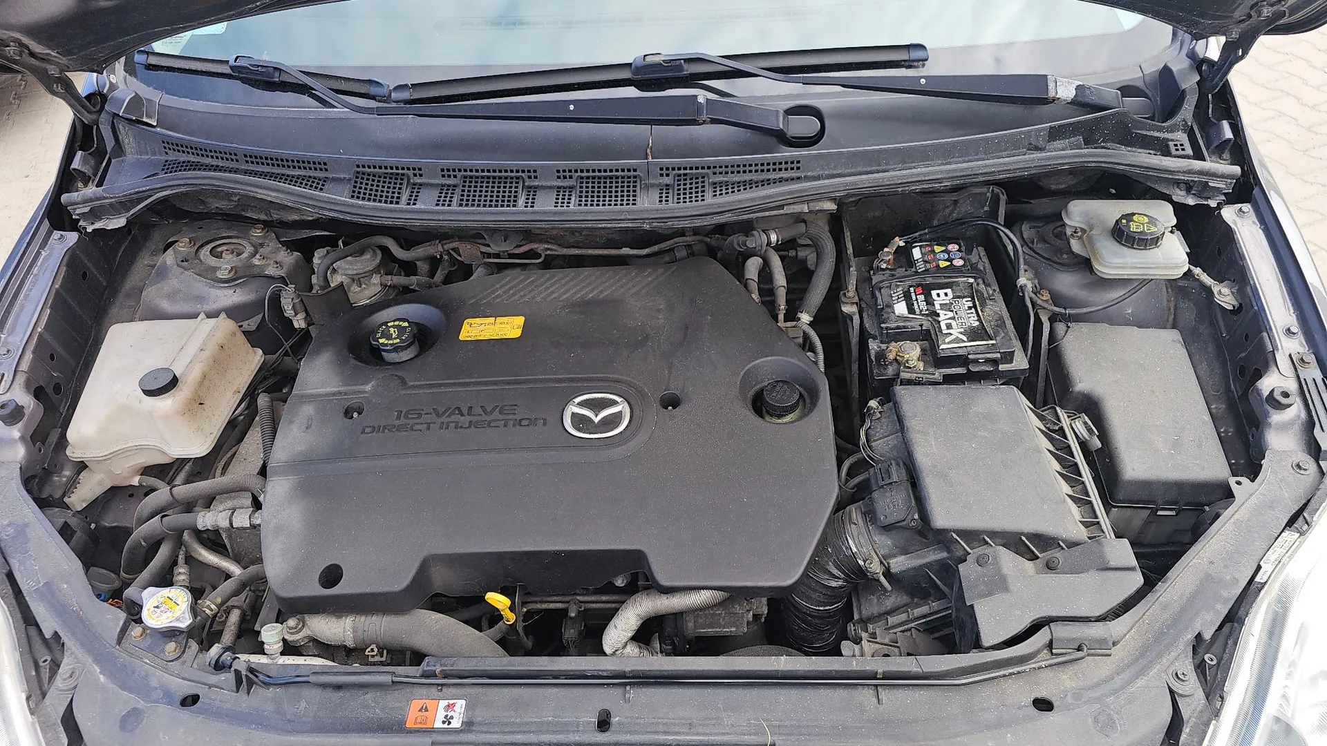Widok z góry na komorę silnika samochodu marki Mazda. W centrum znajduje się czarna plastikowa osłona silnika z napisem '16-VALVE DIRECT INJECTION' oraz logo Mazdy. Po prawej stronie widoczny jest akumulator marki Black Energy oraz czarna obudowa filtra powietrza. Po lewej stronie znajduje się biały zbiornik z płynem. W górnej części zdjęcia widoczna jest przednia szyba z wycieraczkami.