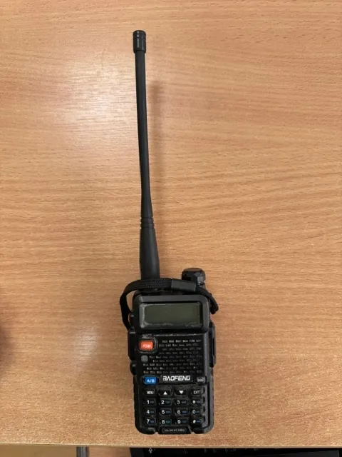 Czarna radiostacja marki Baofeng UV-5R z długą anteną, leżąca na drewnianym blacie. Urządzenie posiada mały ekran LCD, klawiaturę numeryczną oraz pomarańczowy przycisk. Na obudowie widoczny jest napis 'BAOFENG'.