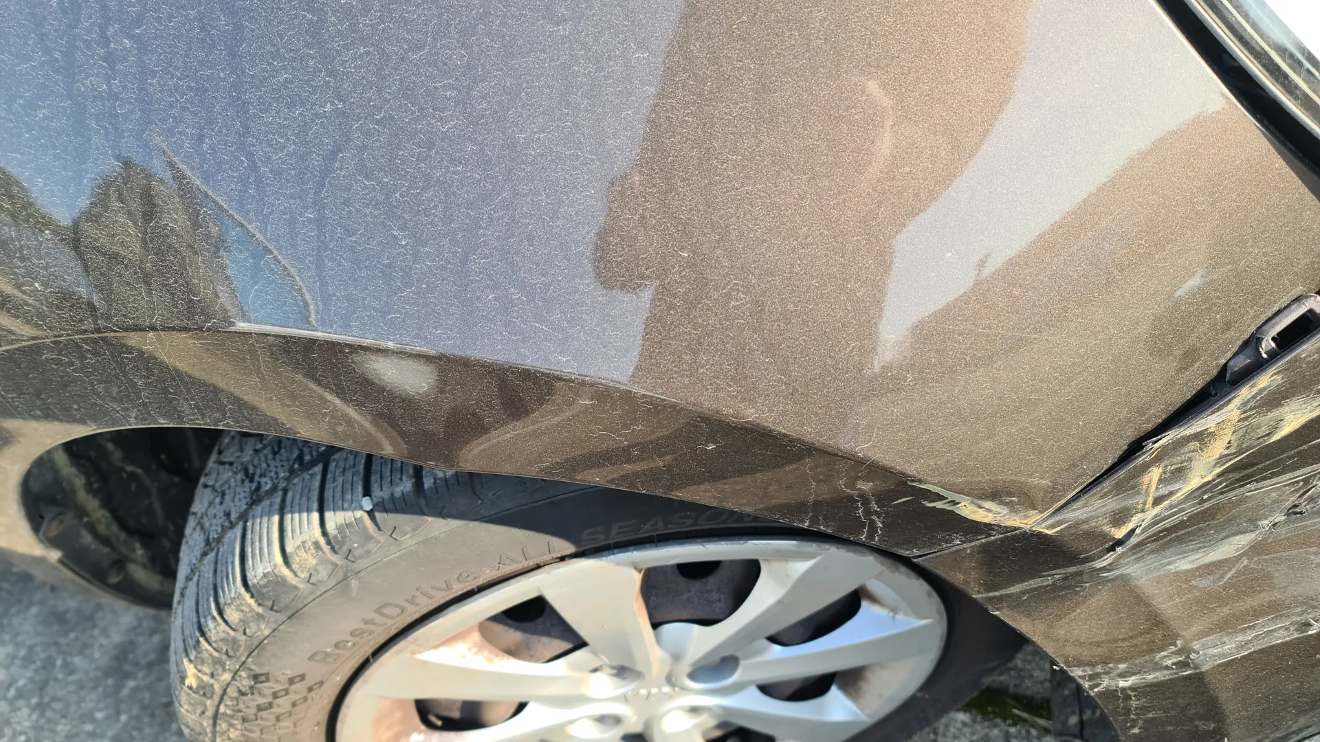 Zbliżenie na przednią prawą część ciemnoszarego samochodu. Widoczne są uszkodzenia na błotniku w postaci zadrapań i pęknięć. Opona z napisem BestDrive All Season oraz srebrna felga z pięcioma promieniami. Na karoserii odbicie osoby trzymającej urządzenie do robienia zdjęć. Tło stanowi asfalt i fragment trawy.