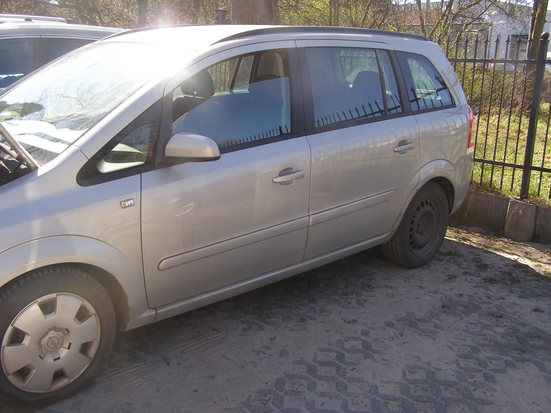Srebrny samochód typu minivan zaparkowany na brukowanej powierzchni. Po lewej stronie widoczna część innego pojazdu. W tle czarny metalowy płot, drzewa oraz budynki. Słońce oświetla przednią część pojazdu, powodując połysk na przedniej szybie. Samochód ma czarne felgi, lusterka boczne i czarne listwy wokół okien. Na drzwiach widoczny mały emblemat. Podłoga składa się z geometrycznego wzoru brukowych płyt.