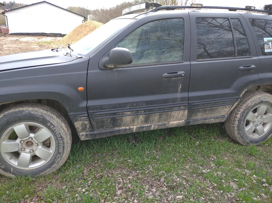 Ciemnoszary SUV marki Jeep Grand Cherokee z widocznym napisem na boku, zaparkowany na trawie z brudnymi oponami i zabrudzonym nadwoziem. W tle biały budynek z czarnym dachem, kopiec ziemi oraz drzewa. Na oponie widoczny napis BFGoodrich All-Terrain T/A. Samochód wyposażony w dachówkę i ma naklejkę na tylnej szybie.