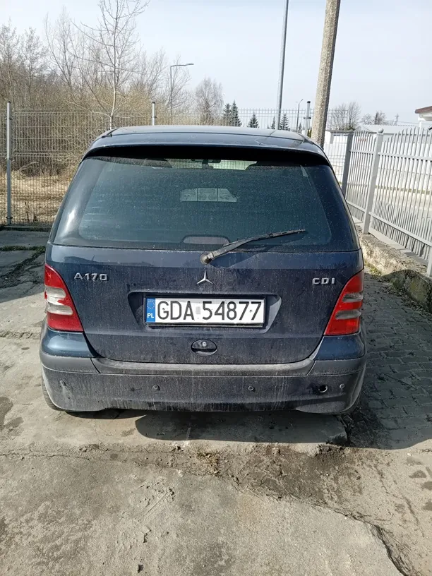 Zdjęcie przedstawia tył ciemnoniebieskiego samochodu Mercedes-Benz klasy A (model A170 CDI). Na bagażniku widoczne są oznaczenia "A170" po lewej stronie i "CDI" po prawej, a w centrum logo marki. Tablica rejestracyjna z polskim kodem "PL" i numerem "GDA 54877" jest umieszczona pionowo na środku. Samochód jest zabrudzony, a na tle widoczne są metalowe ogrodzenia, suche drzewa, słup i betonowa nawierzchnia. W tylnej szybie odbija się niewyraźny obiekt.