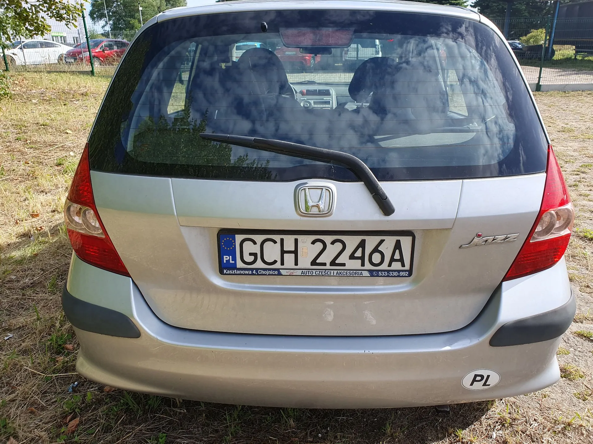 Tylny widok srebrnego samochodu marki Honda Jazz. Na klapie bagażnika znajduje się logo Hondy oraz tablica rejestracyjna o numerze GCH 2246A z oznaczeniem PL. Poniżej tablicy widnieje napis: 'Kasztanowa 4, Chojnice AUTO CZĘŚCI I AKCESORIA 533-330-992'. W prawym dolnym rogu zderzaka znajduje się owalna naklejka PL.