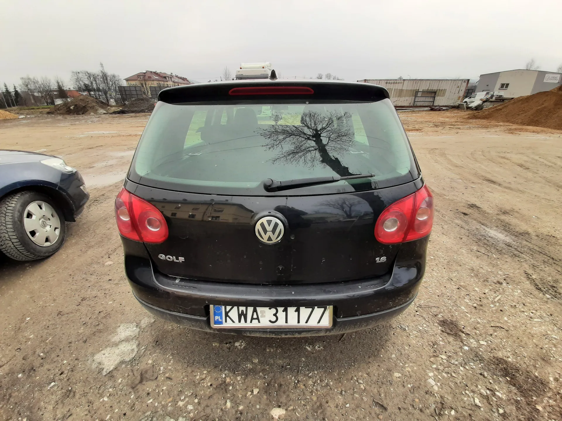 Tył czarnego samochodu Volkswagen Golf z widocznym logo, napisem "GOLF" i oznaczeniem "1.6". Tablica rejestracyjna PL KWA 31177. Samochód stoi na brudnej, żwirowej powierzchni. W tle widoczne są budynki, kopce ziemi, częściowo widoczny niebieski samochód po lewej stronie oraz odbicie drzewa w tylnej szybie. Niebo jest zachmurzone.