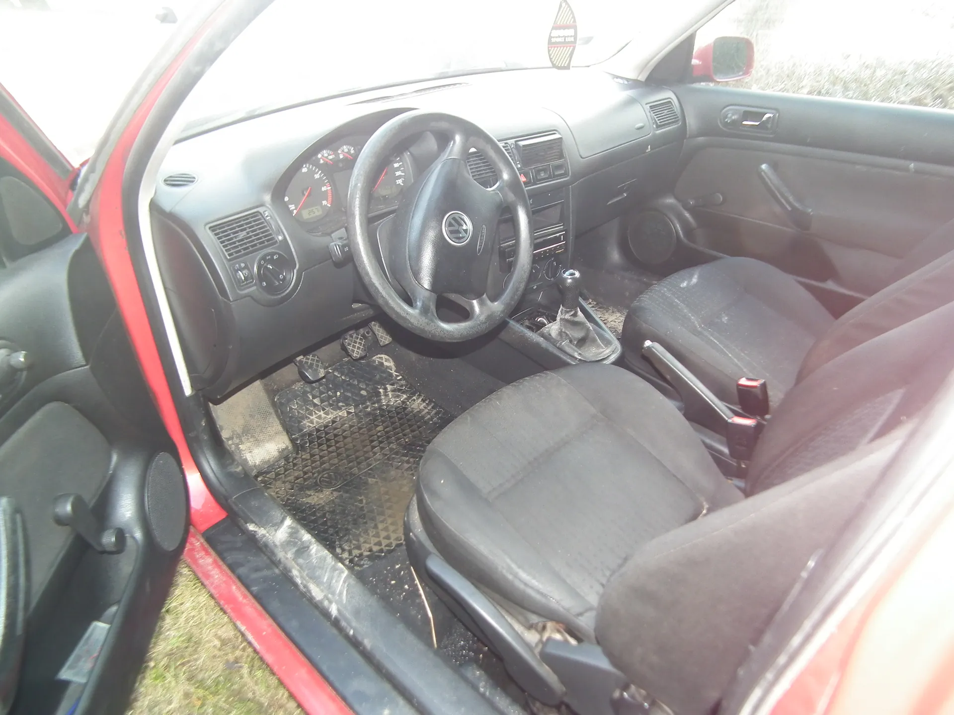 Wnętrze czerwonego samochodu z otwartą drzwiami kierowcy. Widoczna czarna kierownica z logo Volkswagen, deska rozdzielcza z licznikami, czarne siedzenia, brudne dywaniki podłogowe, dźwignia zmiany biegów oraz zewnętrzne lusterko boczne. Poza samochodem widoczna trawa.