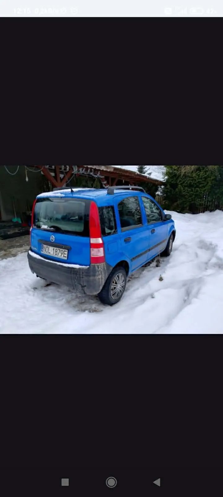 Niebieski samochód osobowy zaparkowany na śniegu, widok od tyłu. Na tablicy rejestracyjnej widoczny napis WOL 1879E. Samochód ma dachowy bagażnik i zabrudzony tylny zderzak. W tle drewniana konstrukcja, drzewa oraz śnieżna powierzchnia z widocznymi odciskami opon.