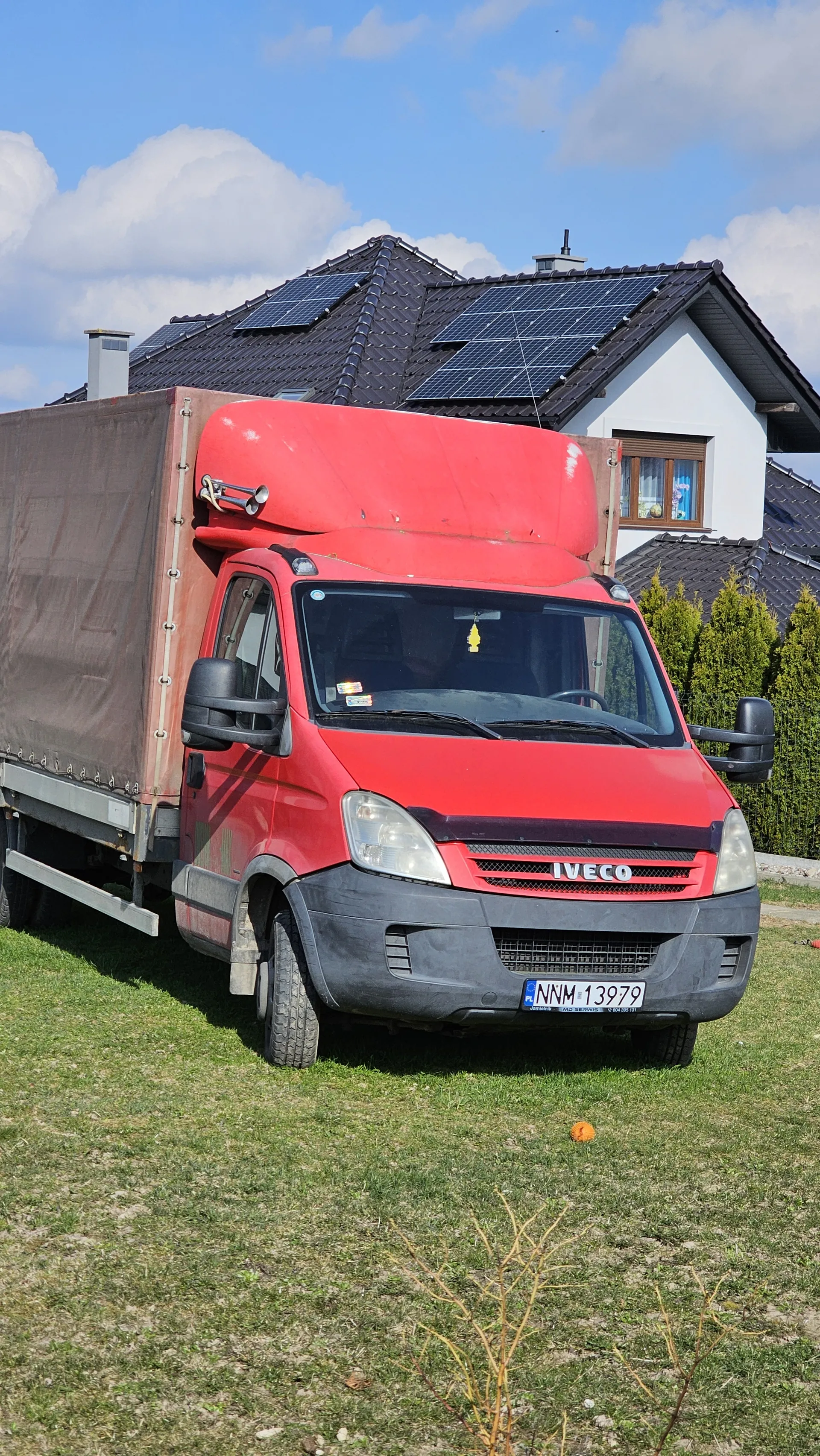 Czerwony samochód dostawczy Iveco z brązowym przyczepą zaparkowany na trawie. Widoczna tablica rejestracyjna NNM 13979. W tle biały dom z czarnym dachem i panelami słonecznymi, zielone krzewy oraz niebo z chmurami.