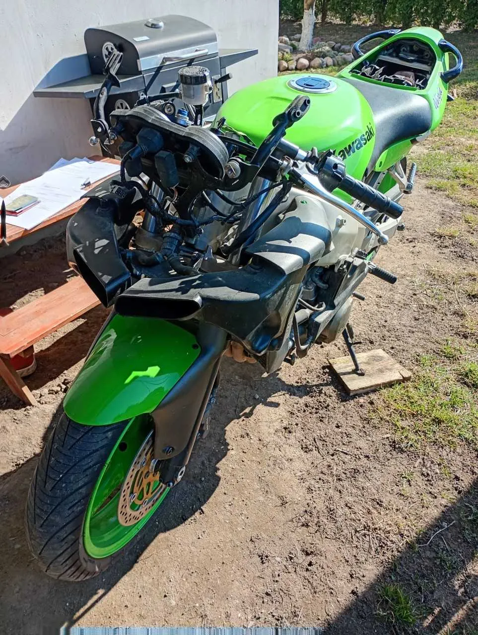 Zdjęcie zielonego motocykla marki Kawasaki zaparkowanego na podwórku. Widoczny zielony zbiornik paliwa z napisem "Kawasaki", czarna siedziba, czarne kierownice i przednie koło z zielonymi akcentami. W tle znajduje się grill, drewniany stół z dokumentami, betonowa ściana oraz trawa z kamieniami. Motocykl stoi na podkładowym desku, a podłoże jest ziemne z fragmentami trawy.