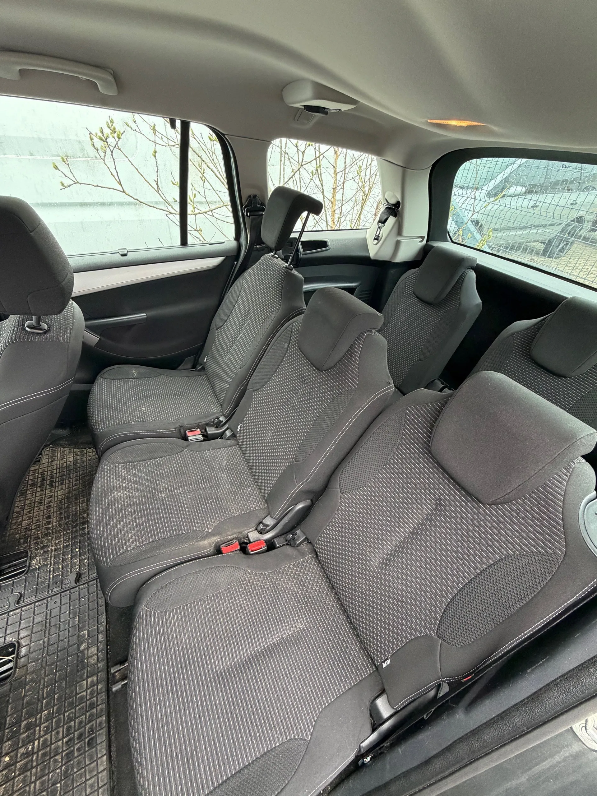 Wnętrze samochodu z widokiem na tylne siedzenia. Siedzenia tapicerowane szarym materiałem z geometrycznym wzorem, z widocznymi pasami bezpieczeństwa z czerwonymi przyciskami. Podłoga wyłożona czarną gumową matą. Przez okna widać drzewa bez liści i budynek na zewnątrz. Sufit samochodu w jasnym kolorze.