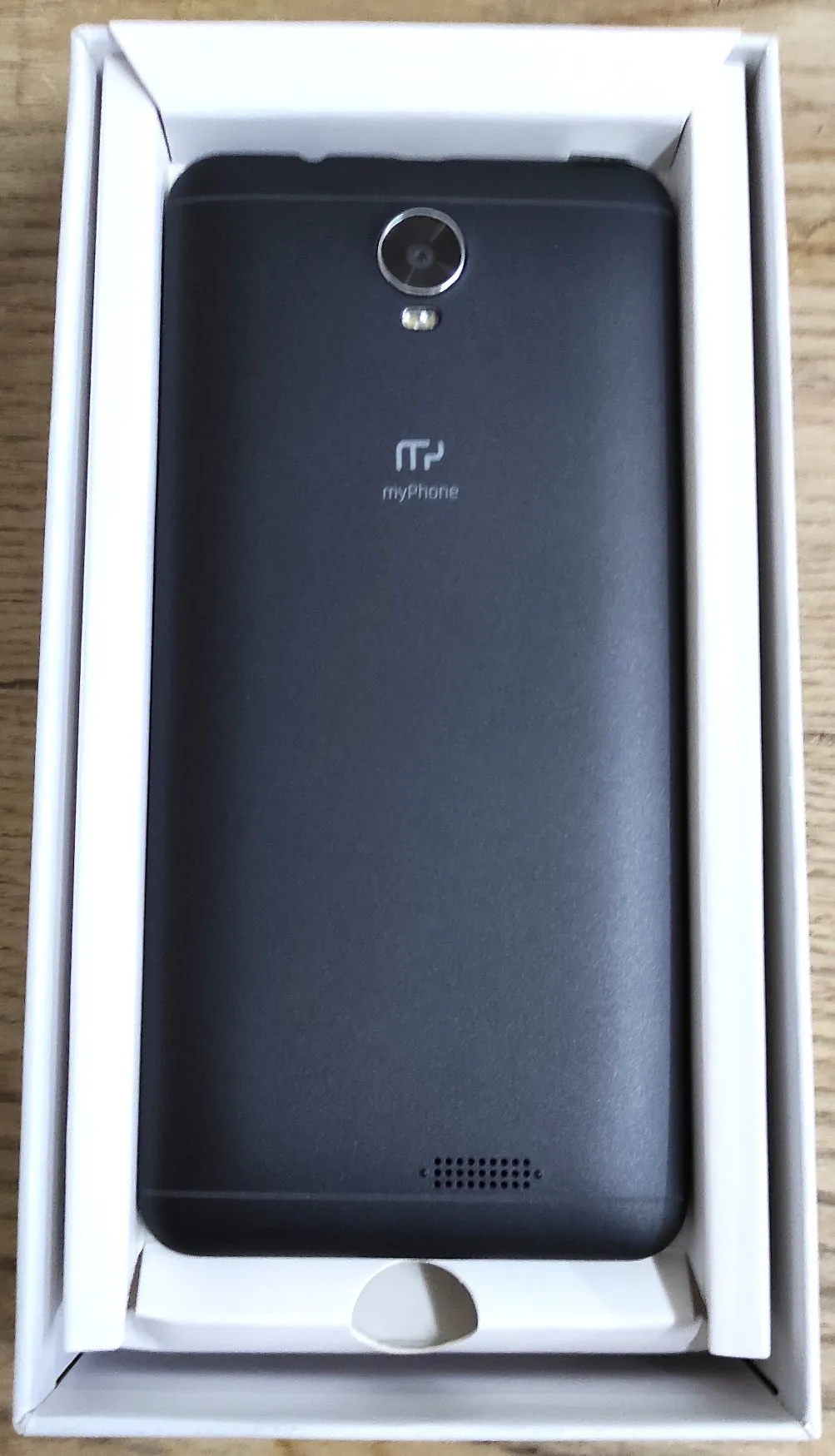 Czarny smartfon marki myPhone leżący w białym, kartonowym opakowaniu. Na tylnej obudowie telefonu widoczny jest okrągły aparat fotograficzny z lampą błyskiem, logo myPhone oraz otwory głośnika na dole.