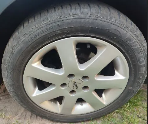 Zbliżenie na koło samochodowe z oponą i srebrną felgą aluminiową o pięciu szerokich ramionach. Na boku opony widoczny jest napis '185/55R15'. W centrum felgi znajduje się mała okrągła nasadka z logo 'AUEC'.