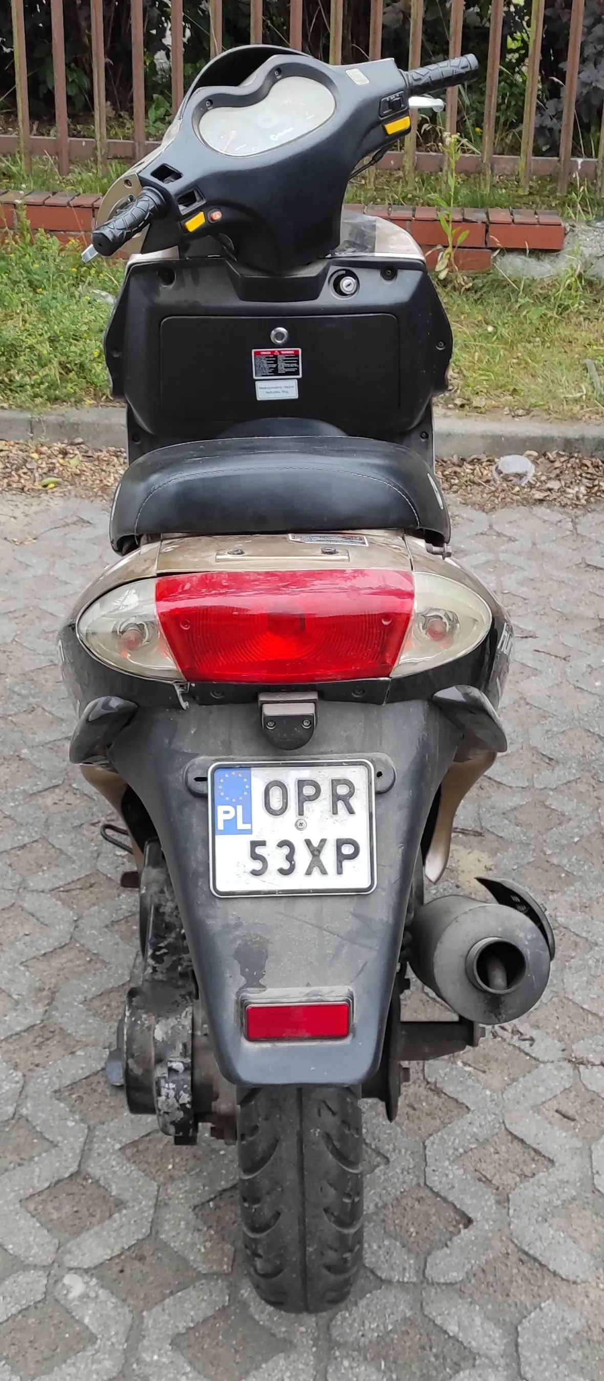 Widok z tyłu na skuter w kolorze złotym i czarnym, zaparkowany na kostce brukowej. Pojazd posiada polską tablicę rejestracyjną o numerze OPR 53XP. Widoczne jest czerwone tylne światło, czarne siedzenie oraz wydech po prawej stronie.