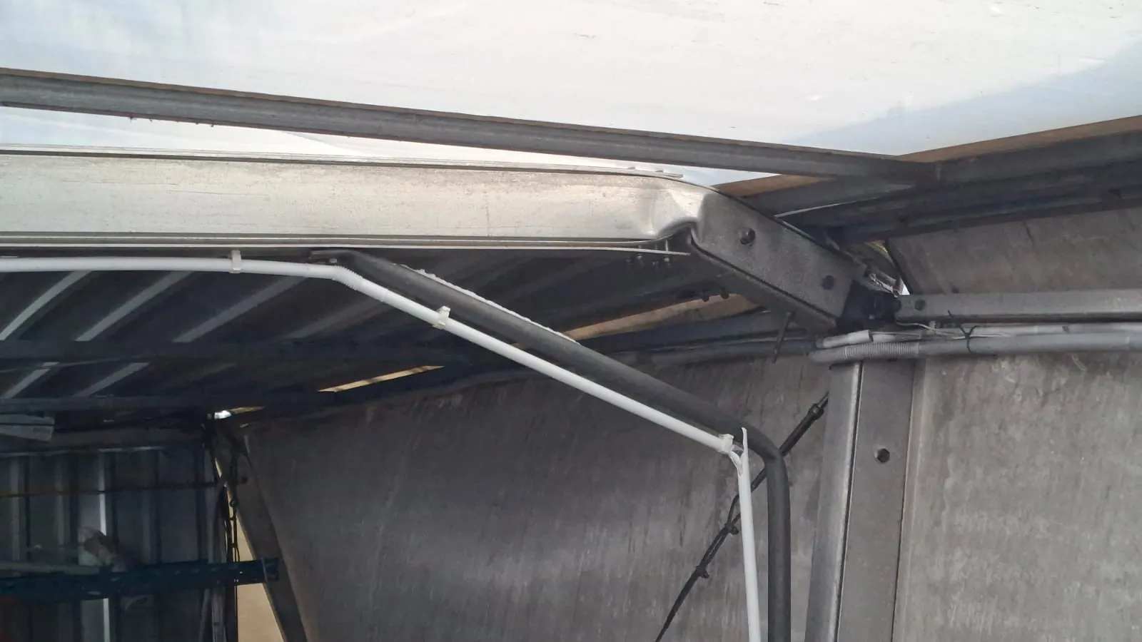 Zdjęcie przedstawia fragment metalowej konstrukcji przemysłowej lub pojazdu. Widoczne są poziome i pionowe elementy metalowe, w tym rury, przewody oraz białe kable przytwierdzone plastikowymi zaciskami. W tle znajduje się powierzchnia betonowa lub metalowa. Brak czytelnego tekstu.