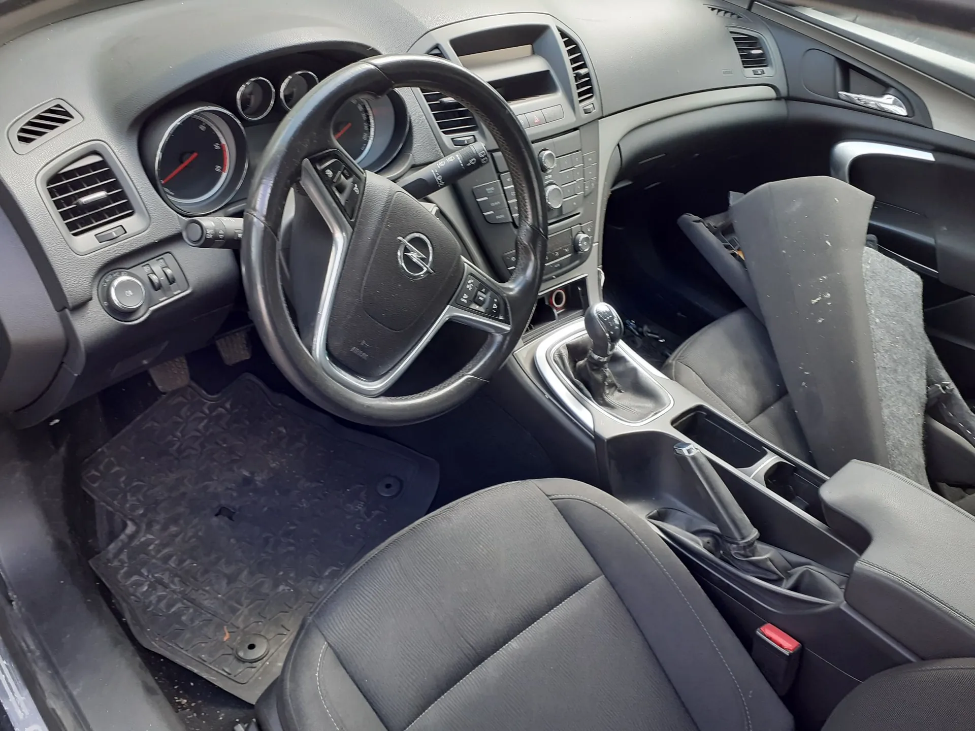 Wnętrze samochodu marki Opel z perspektywy kierowcy. Widoczna jest czarna kierownica z logo producenta, deska rozdzielcza z zegarami, nawiewy oraz konsola środkowa z lewarkiem zmiany biegów i hamulcem ręcznym. Fotele są obite czarnym materiałem, a na podłodze znajduje się gumowa mata. Fotel pasażera jest odchylony do tyłu.